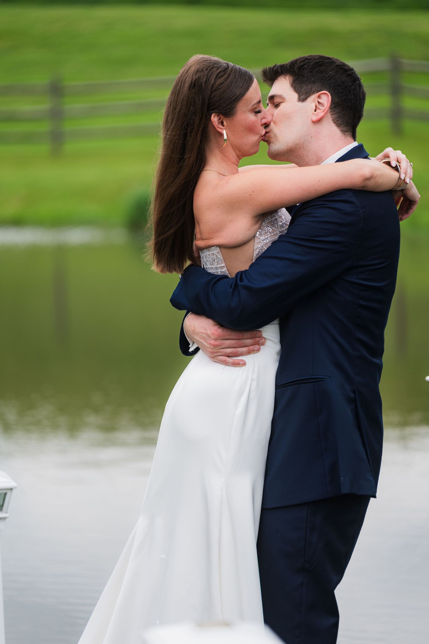 A romantic wedding moment as the couple shares a kiss beside a pond with a rustic fence in the background.