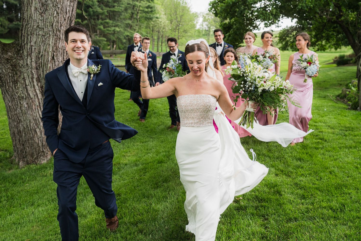 A newlywed couple dances playfully on grass while their wedding party watches behind them.  at shady elms farm pittsburgh pa photography an