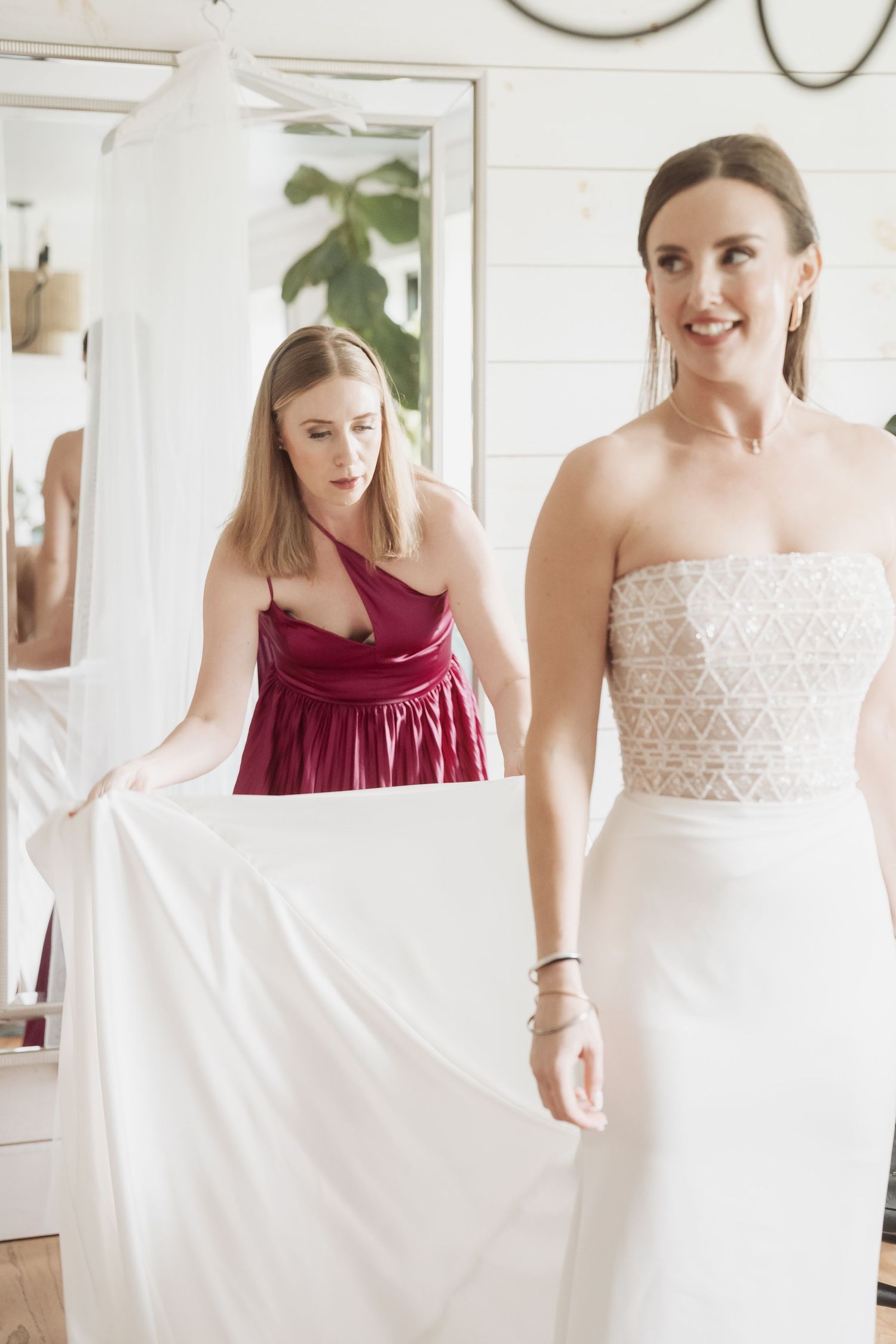 A bride in a white lace gown getting dressed with help from an attendant in burgundy.