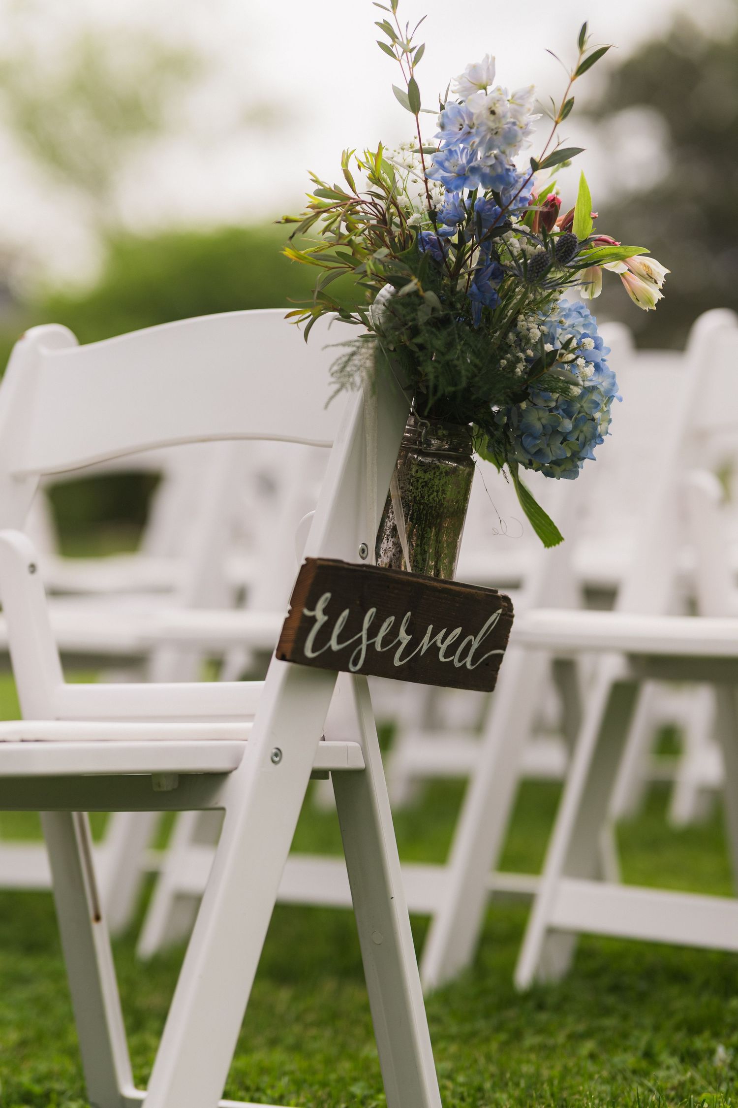 White folding chair with mason jar of blue hydrangeas and reserved sign at outdoor wedding.