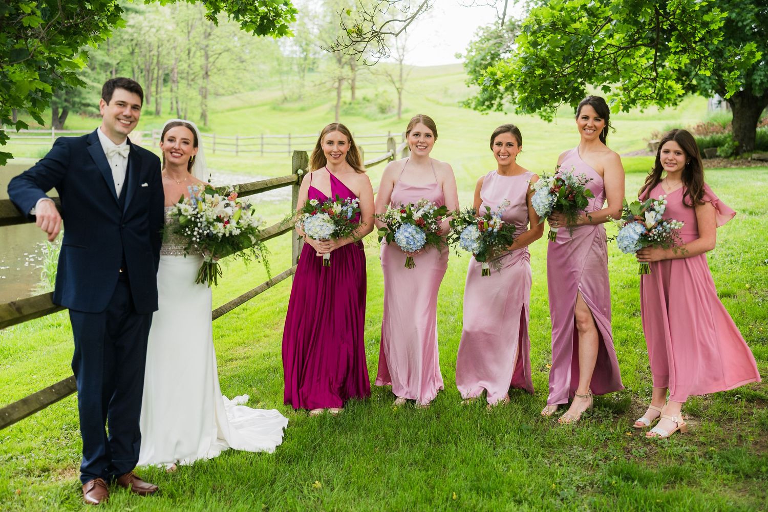 Wedding party in pink and magenta dresses posing outdoors with greenery and rolling hills in background.  at shady elms farm pittsburgh pa