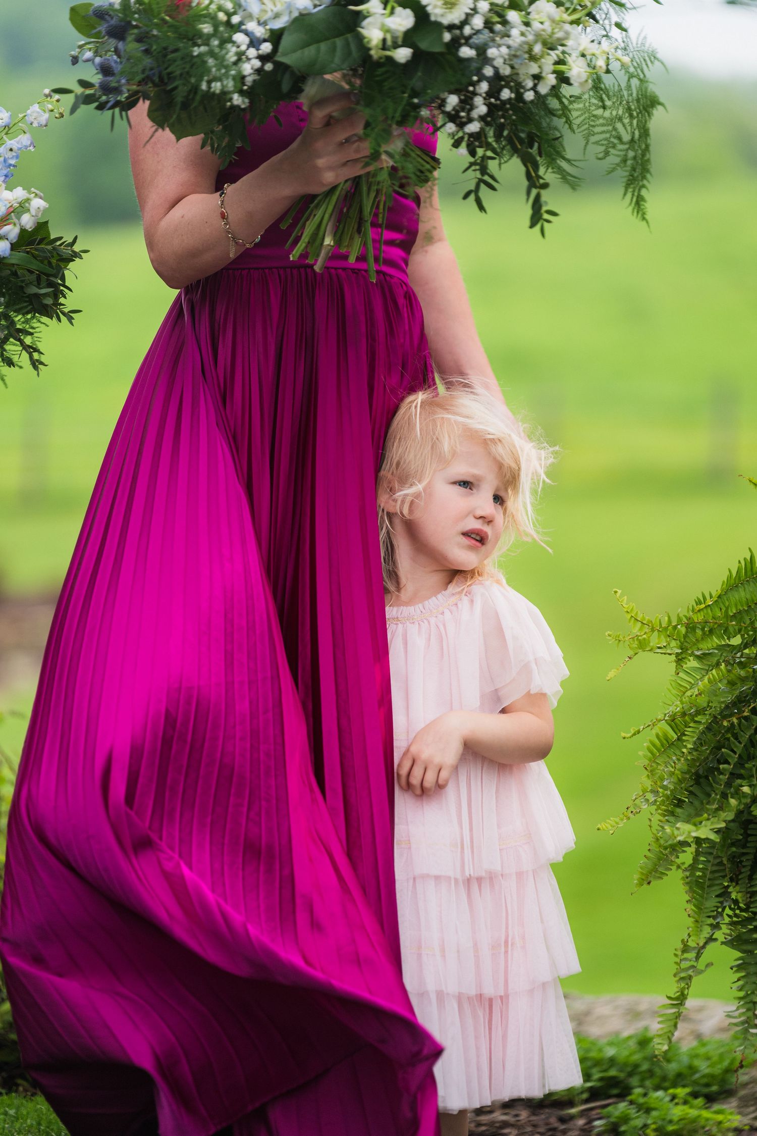A small child in a light pink dress stands beside someone wearing a vibrant magenta gown holding fresh flowers.