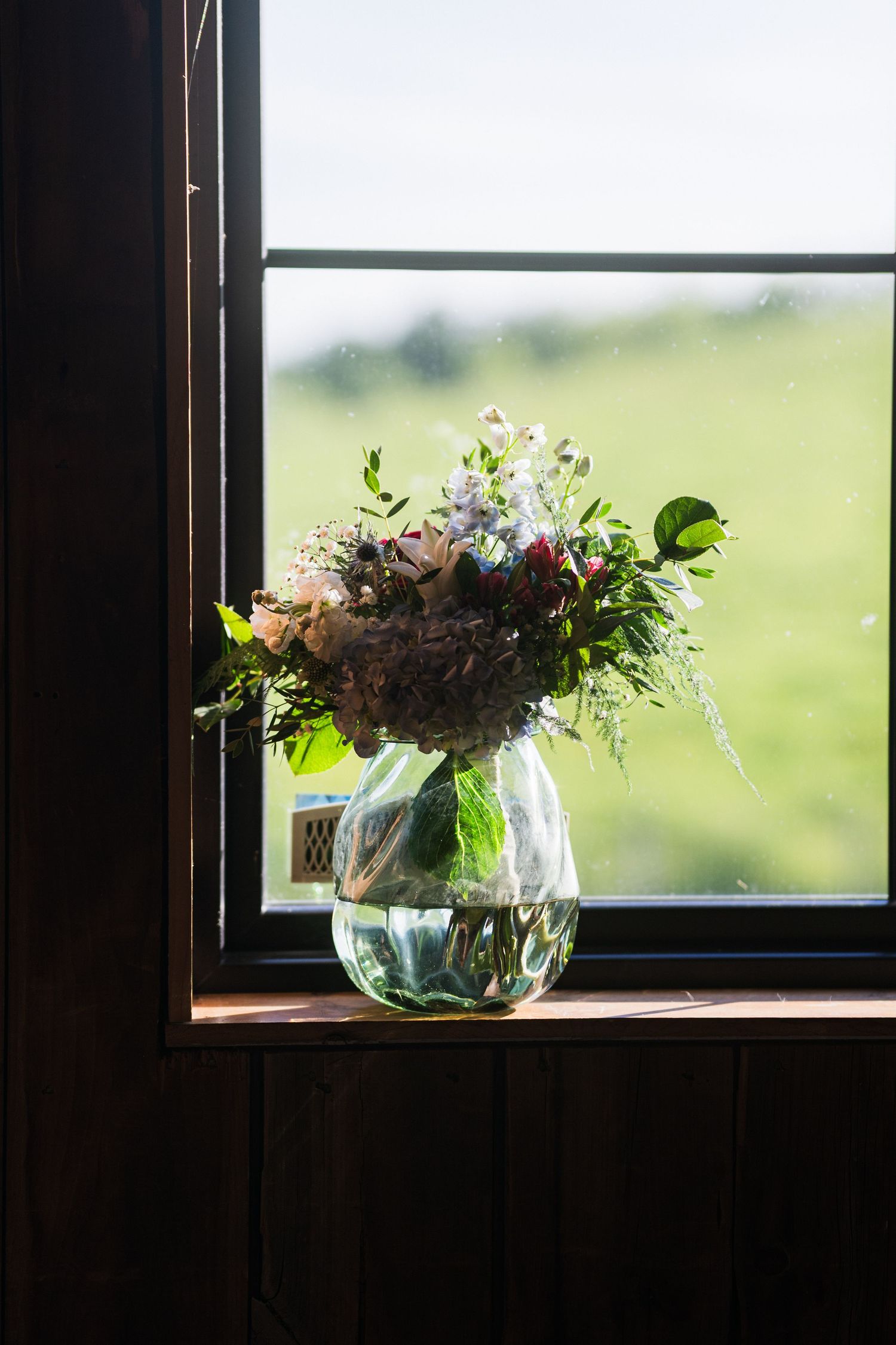Fresh floral arrangement in glass vase sitting on windowsill with bright outdoor view.