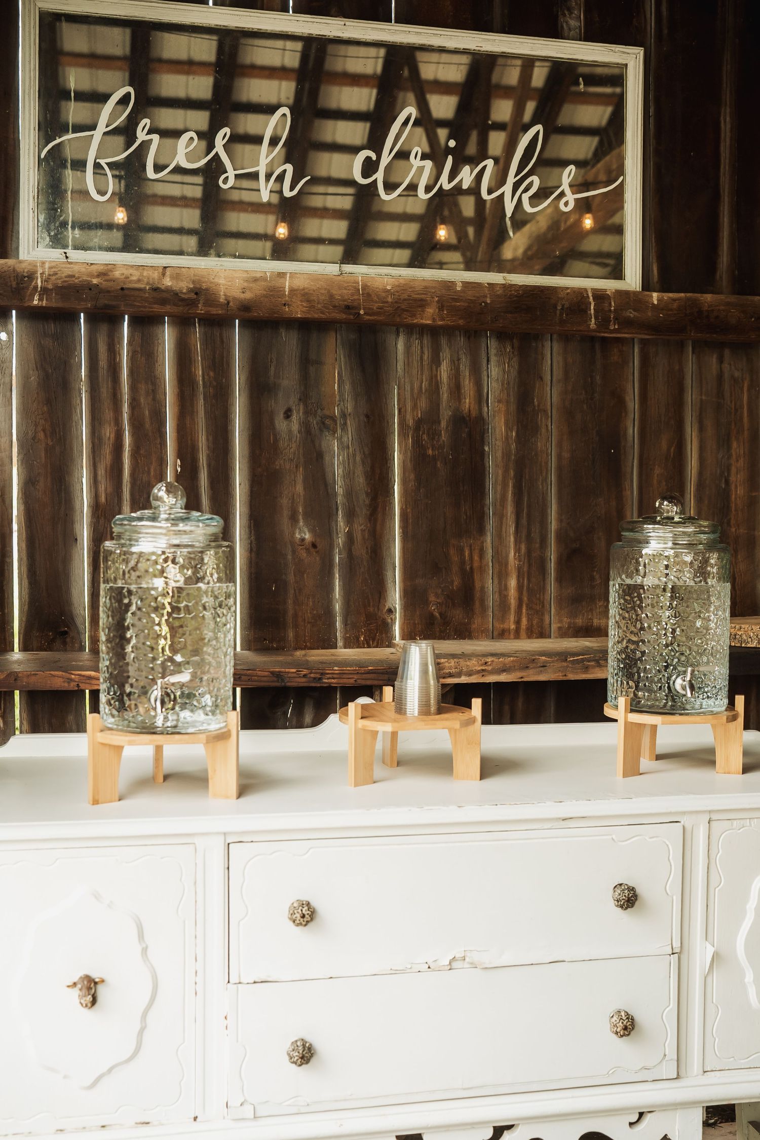 Rustic drink station with glass dispensers on wooden stands under a 'fresh drinks' neon sign and white vintage dresser.