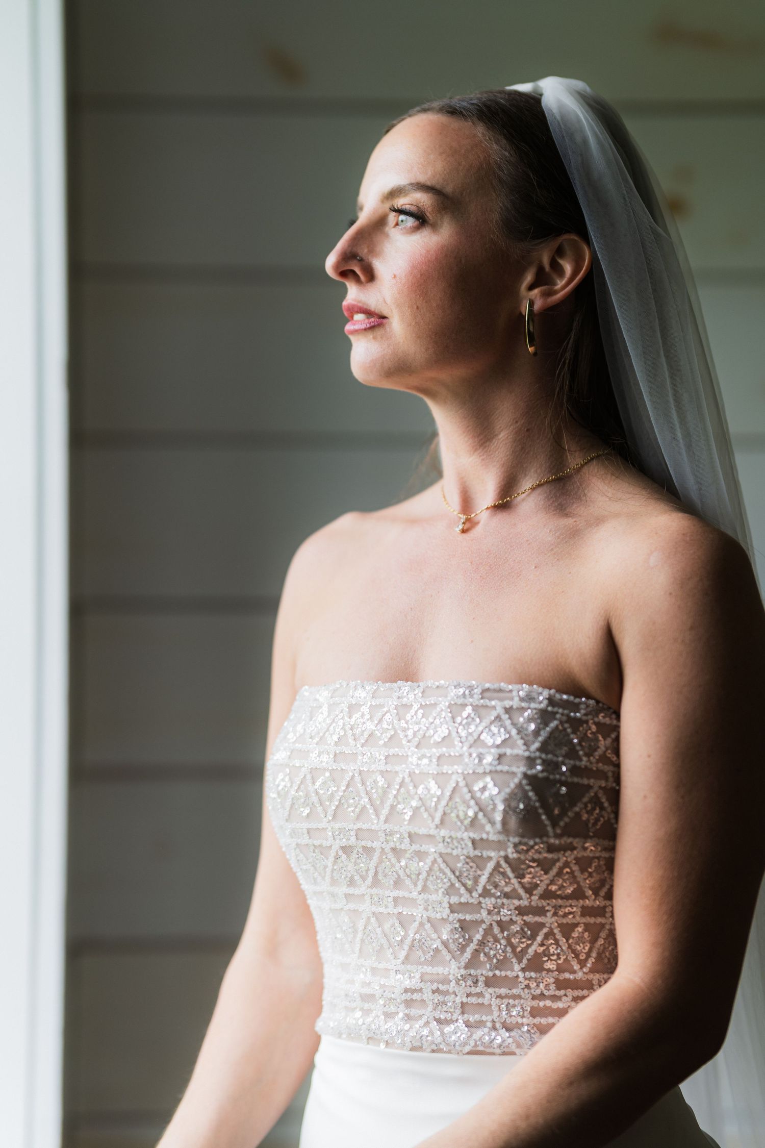 A bride in a strapless lace wedding gown gazes thoughtfully by a window in natural light.  at shady elms farm pittsburgh pa photography and