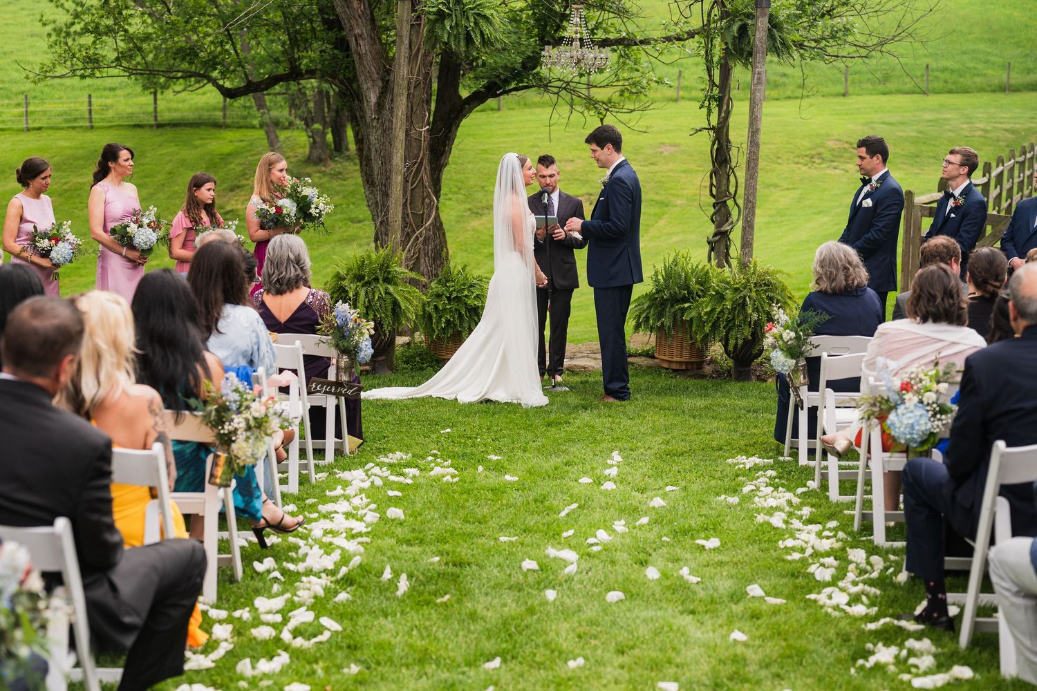 Wedding ceremony in progress under trees with guests seated on white chairs and petals down the aisle.  at shady elms farm pittsburgh pa ph