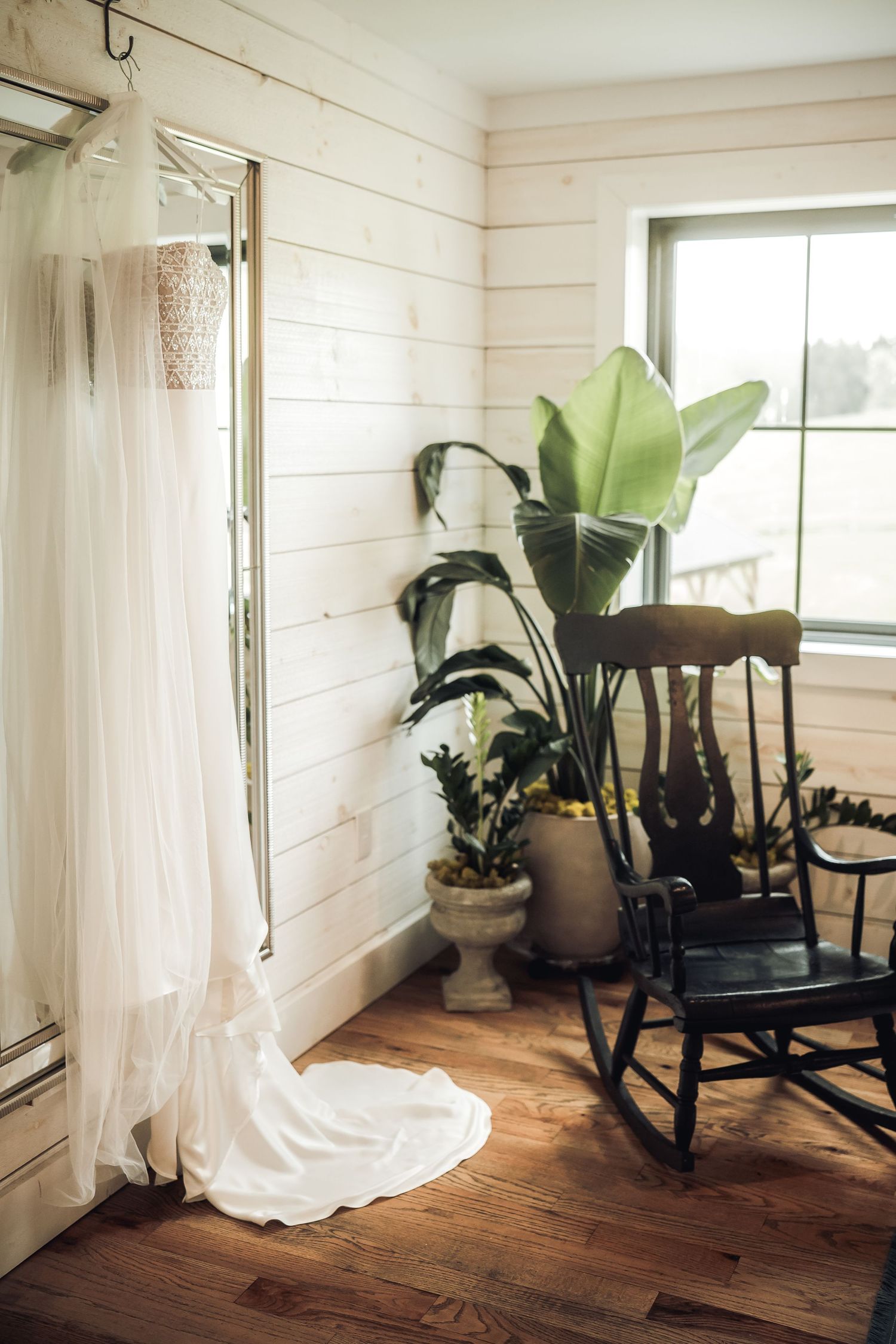 A white wedding dress hangs near a rocking chair and potted plants in a rustic room with shiplap walls.