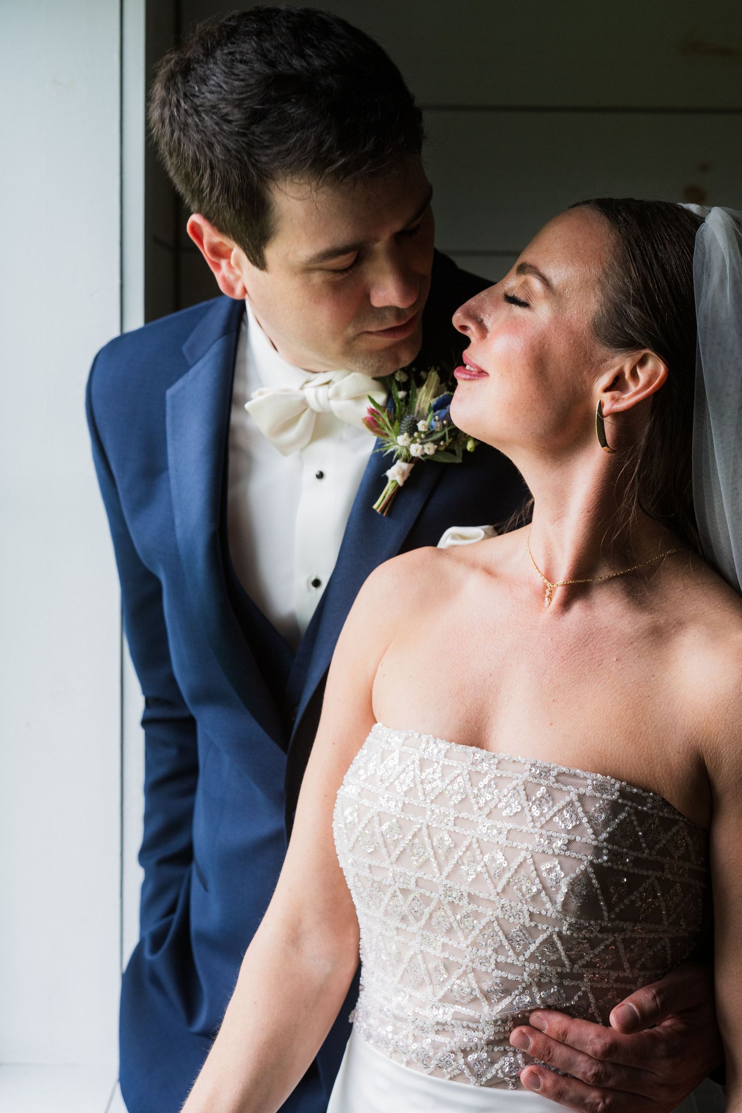 A romantic wedding portrait of a couple in a navy suit and white lace gown sharing an intimate moment by a window  at shady elms farm pitts