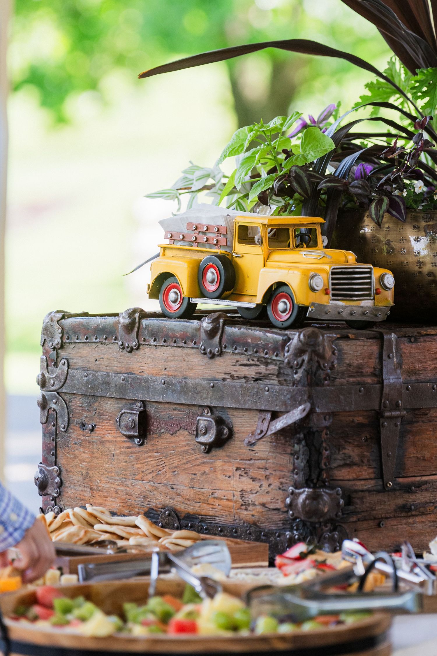 Vintage yellow toy truck displayed on rustic wooden chest with plants and table settings in foreground.
