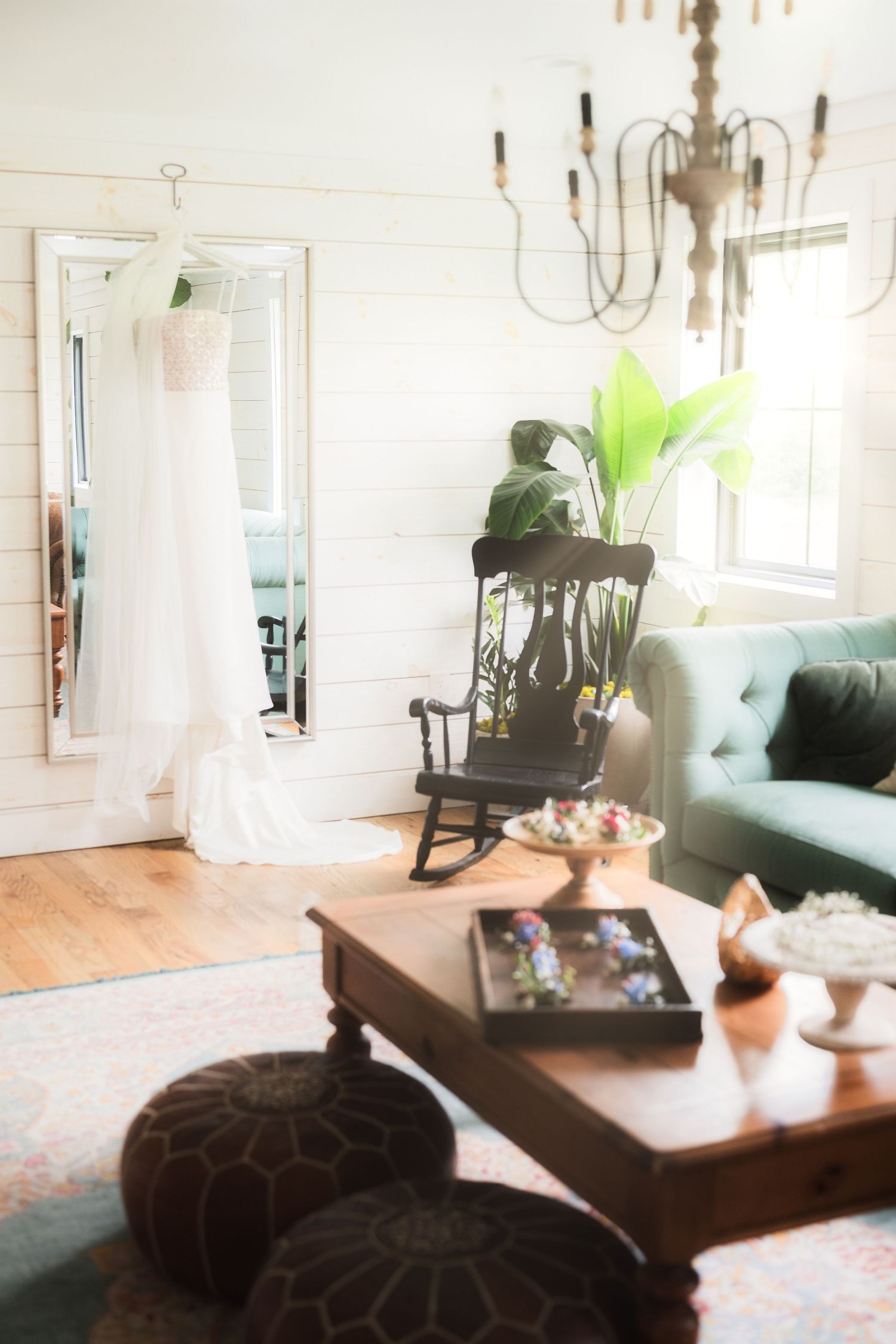 White wedding dress hanging in a cozy room with a vintage rocking chair, chandelier, and teal sofa.