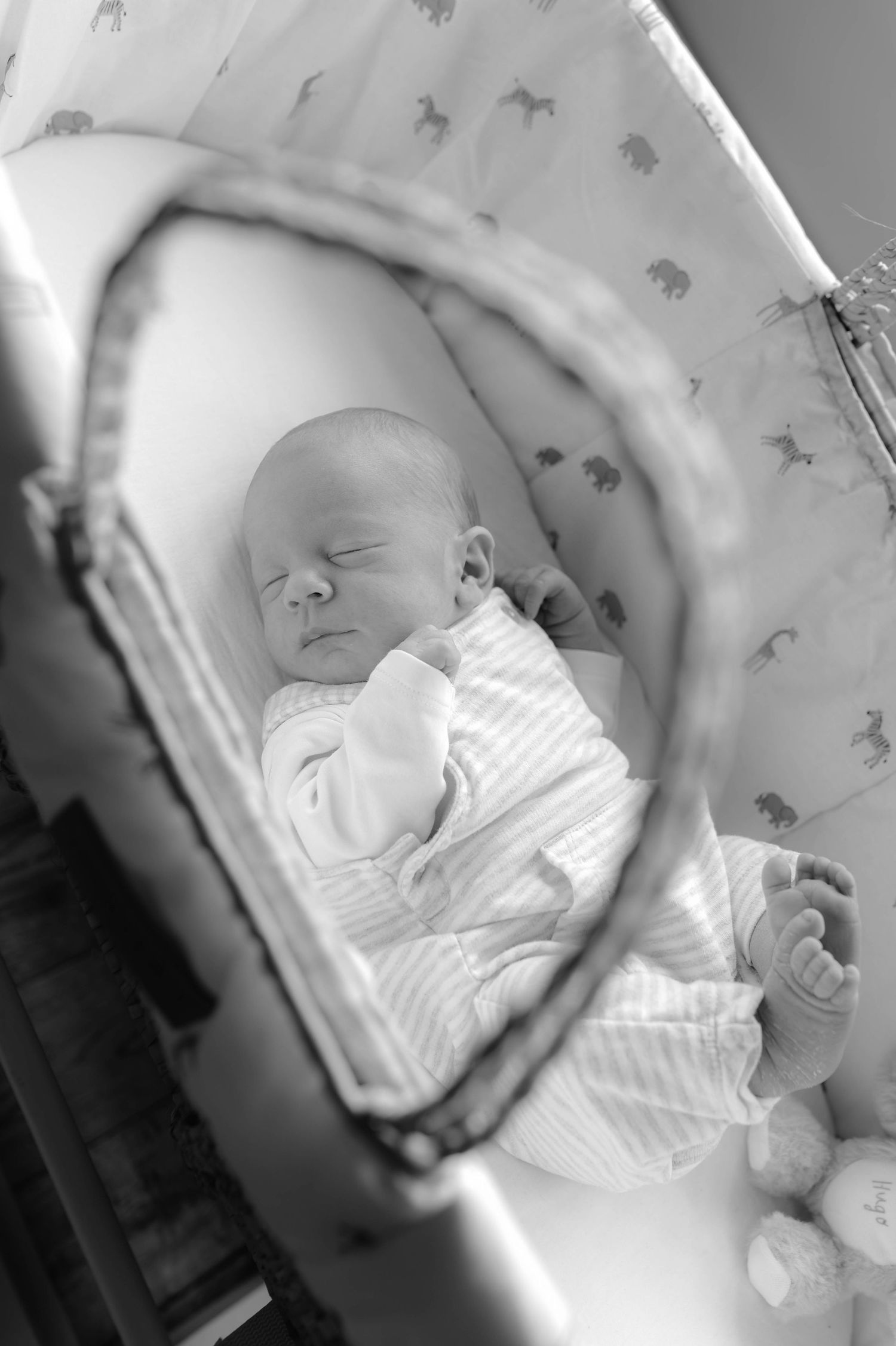 A sleeping newborn baby in white clothing rests peacefully in a patterned bassinet, captured in black and white.