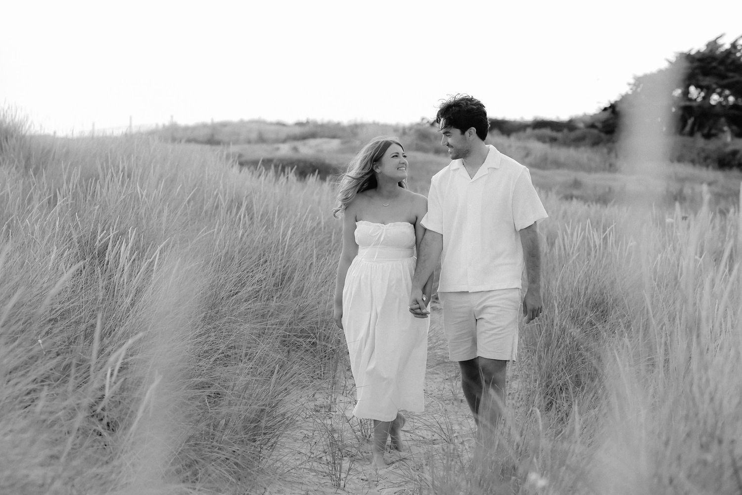 A couple in white outfits holds hands while standing together in tall beach grass on a sunny summer day.