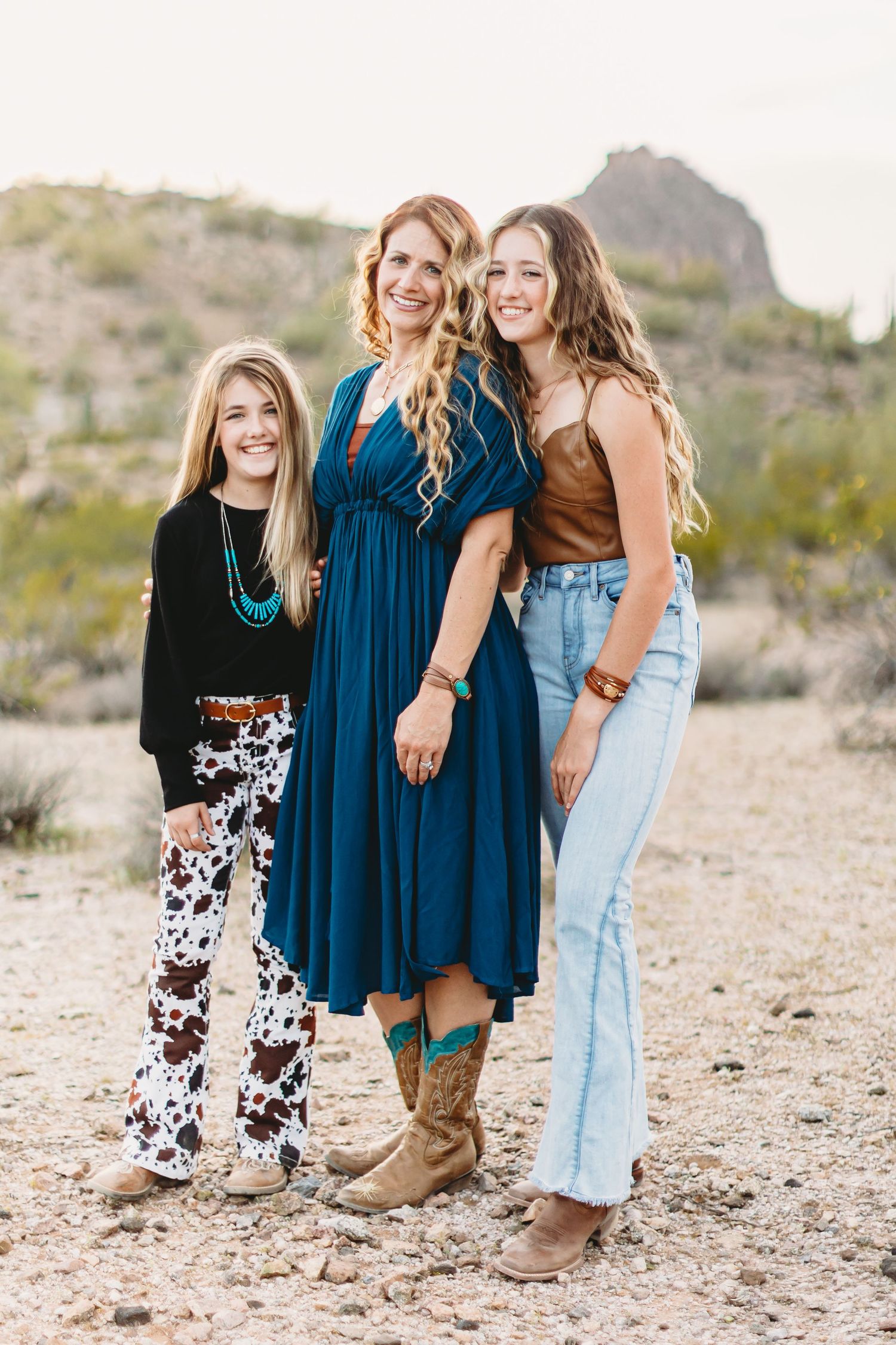 Three friends in casual western outfits stand together on desert terrain with mountains in the background.