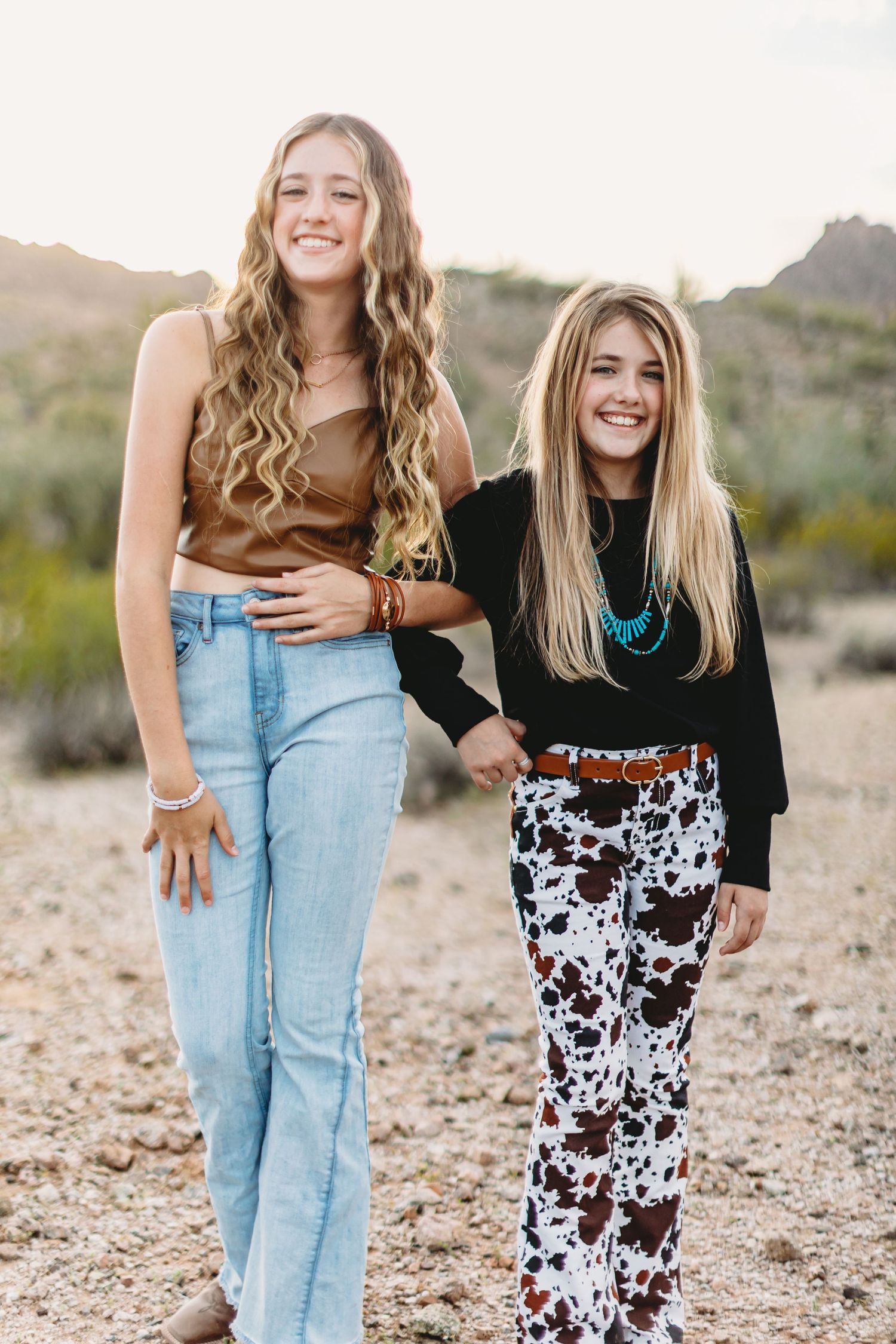 Two friends pose together in casual outfits against a desert landscape during golden hour.