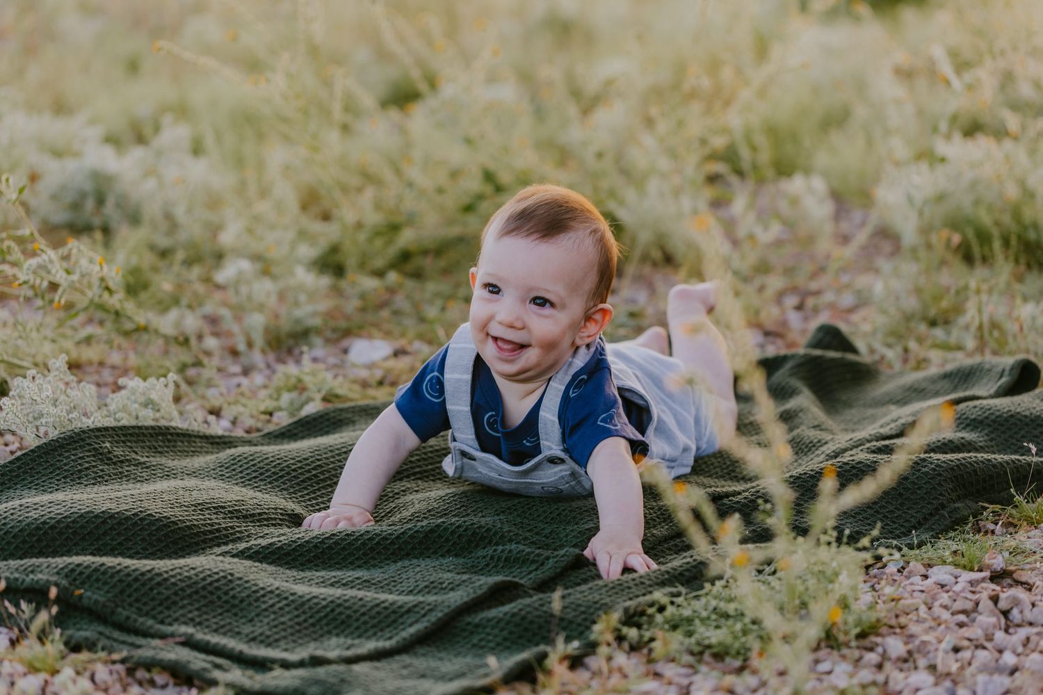 Child plays and rolls on a green blanket outdoors in a sunny field during a summer day.