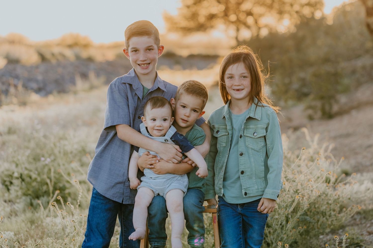 Family wearing coordinated blue and green outfits poses together in a golden desert landscape at sunset.