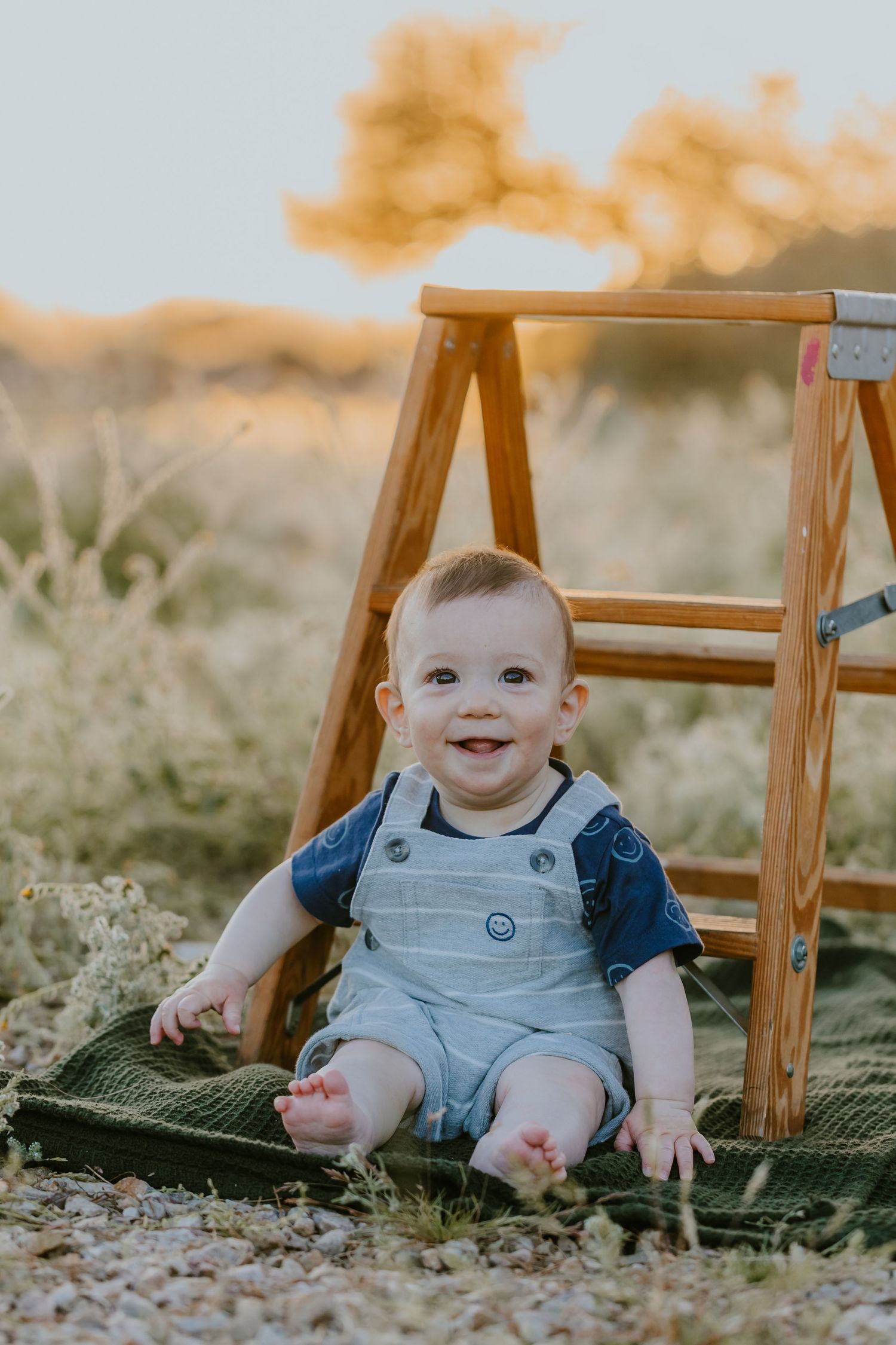 Baby in denim overalls sits on green blanket outdoors during golden sunset light.