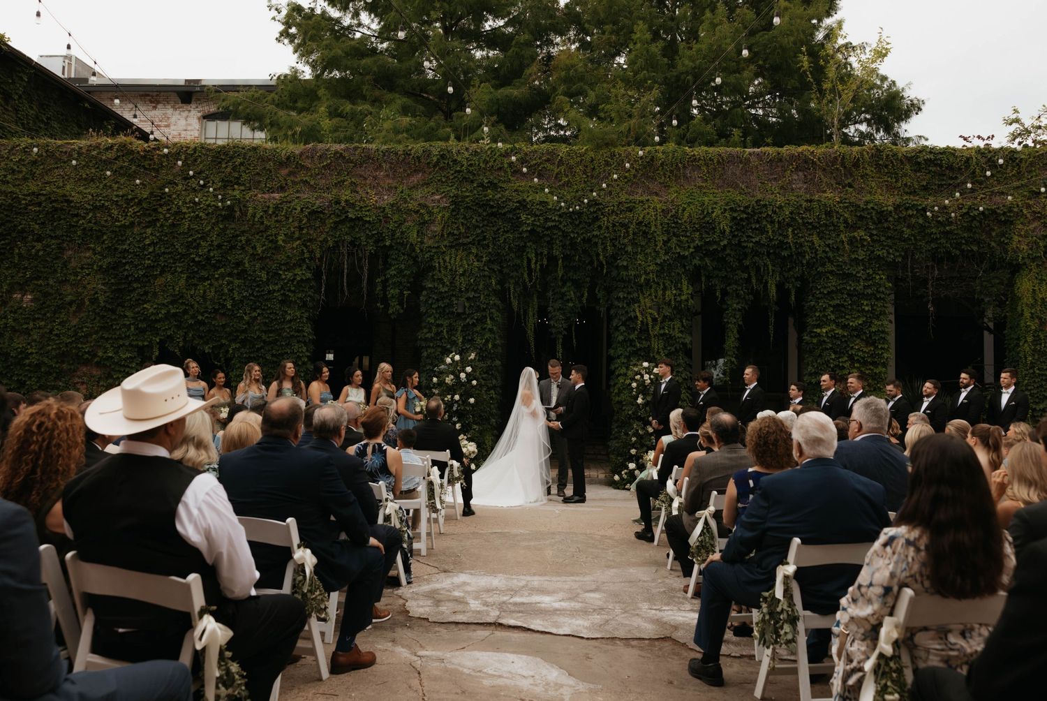 Outdoor wedding ceremony with guests seated in white chairs along an ivy-covered wall aisle, couple at altar.