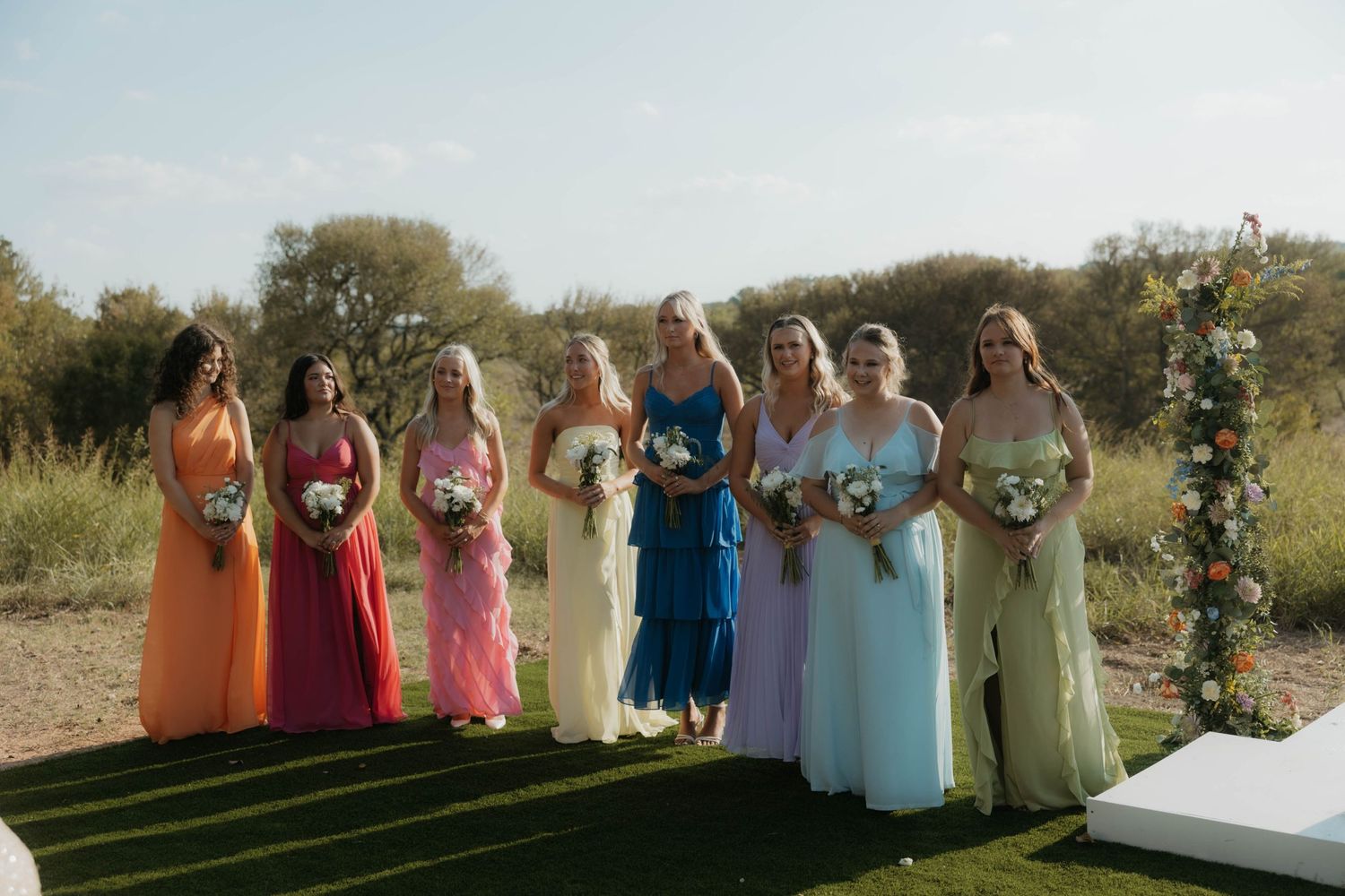 Seven bridesmaids in rainbow colored gowns holding bouquets at outdoor wedding ceremony.
