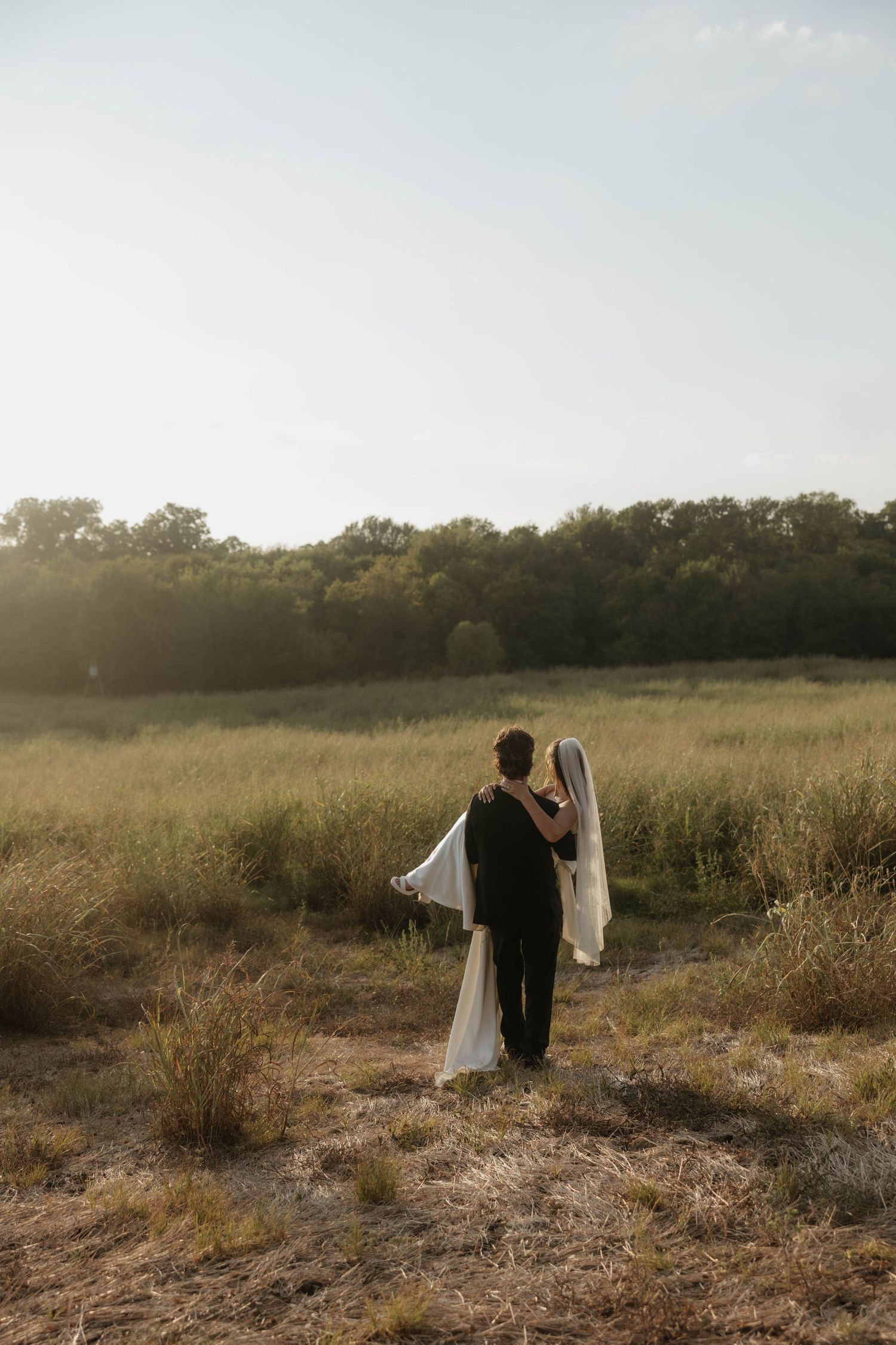 Bride and groom embracing while gazing at golden hour sunset in open field.