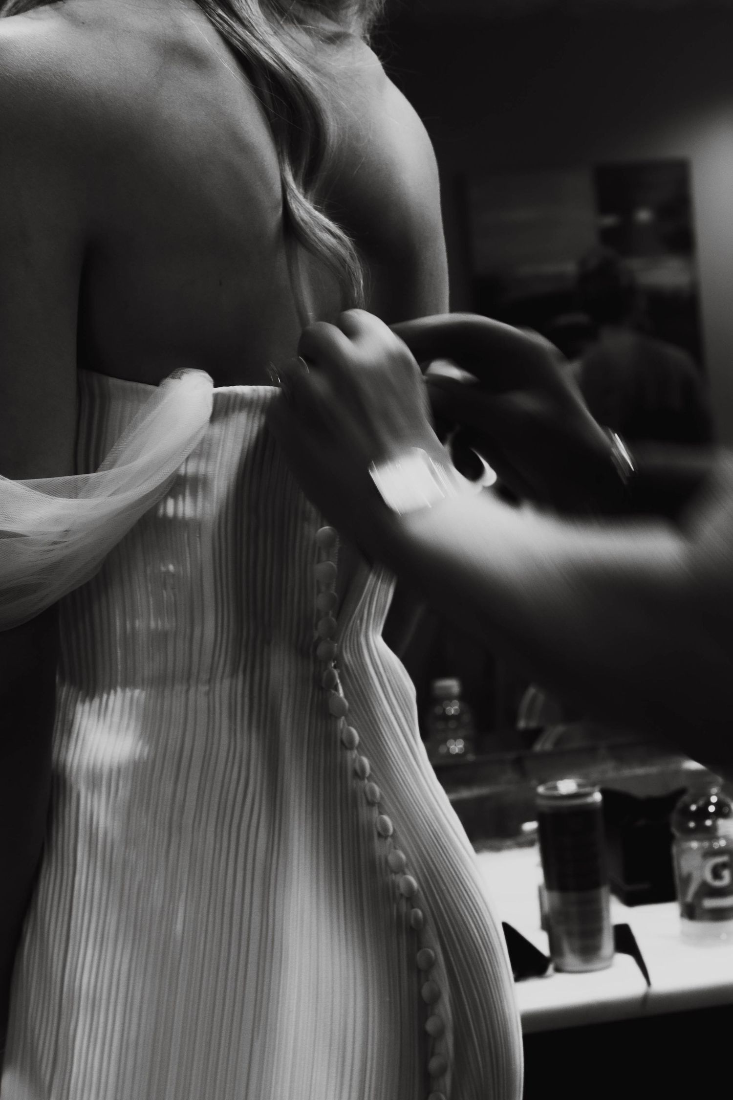 Black and white photo of a woman in a pleated corset dress being buttoned up from behind in a dressing room.