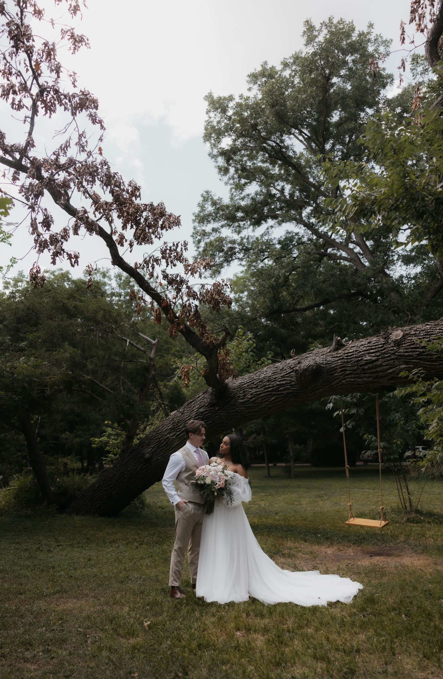 Bride and groom share romantic moment under large fallen oak tree on lush green wedding day.