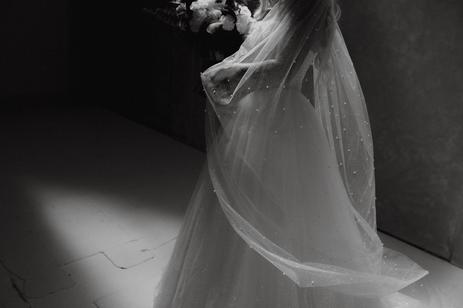 Elegant bride in a flowing wedding gown and veil holding a bouquet in dramatic black and white.