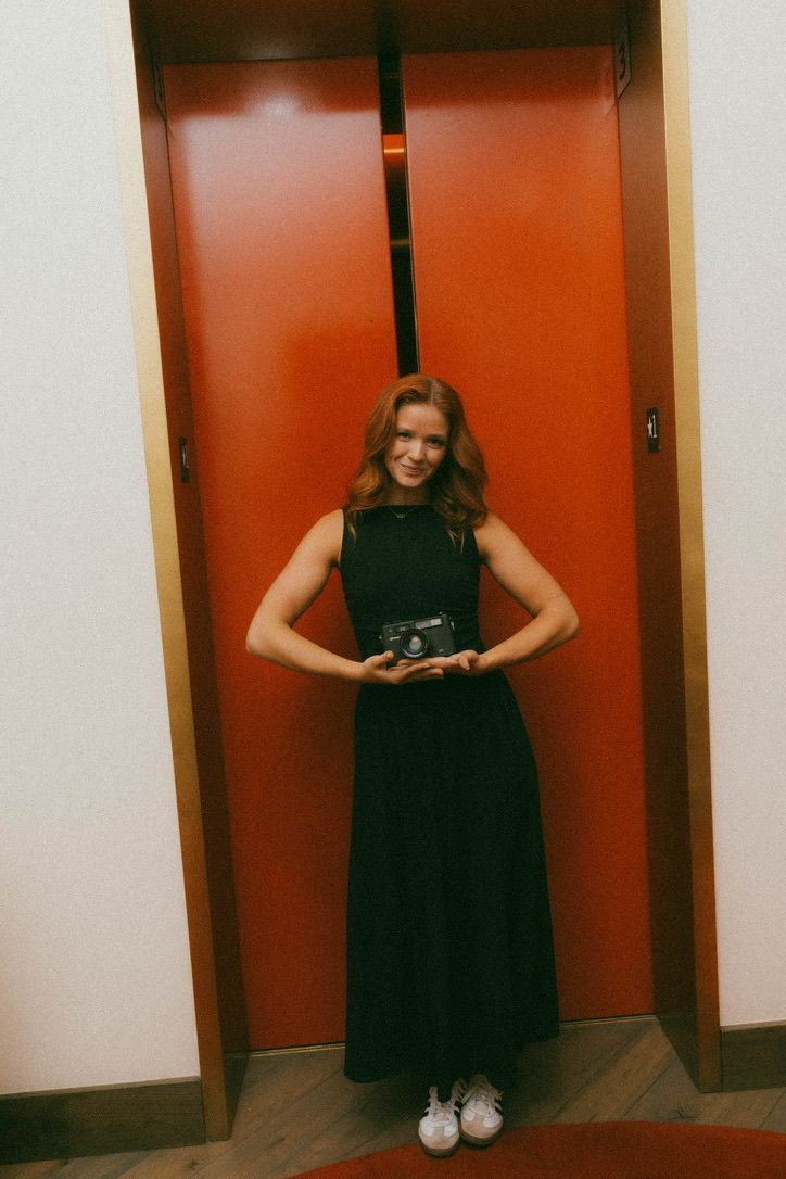 Woman in black maxi dress holding vintage camera, smiling in front of red elevator doors.