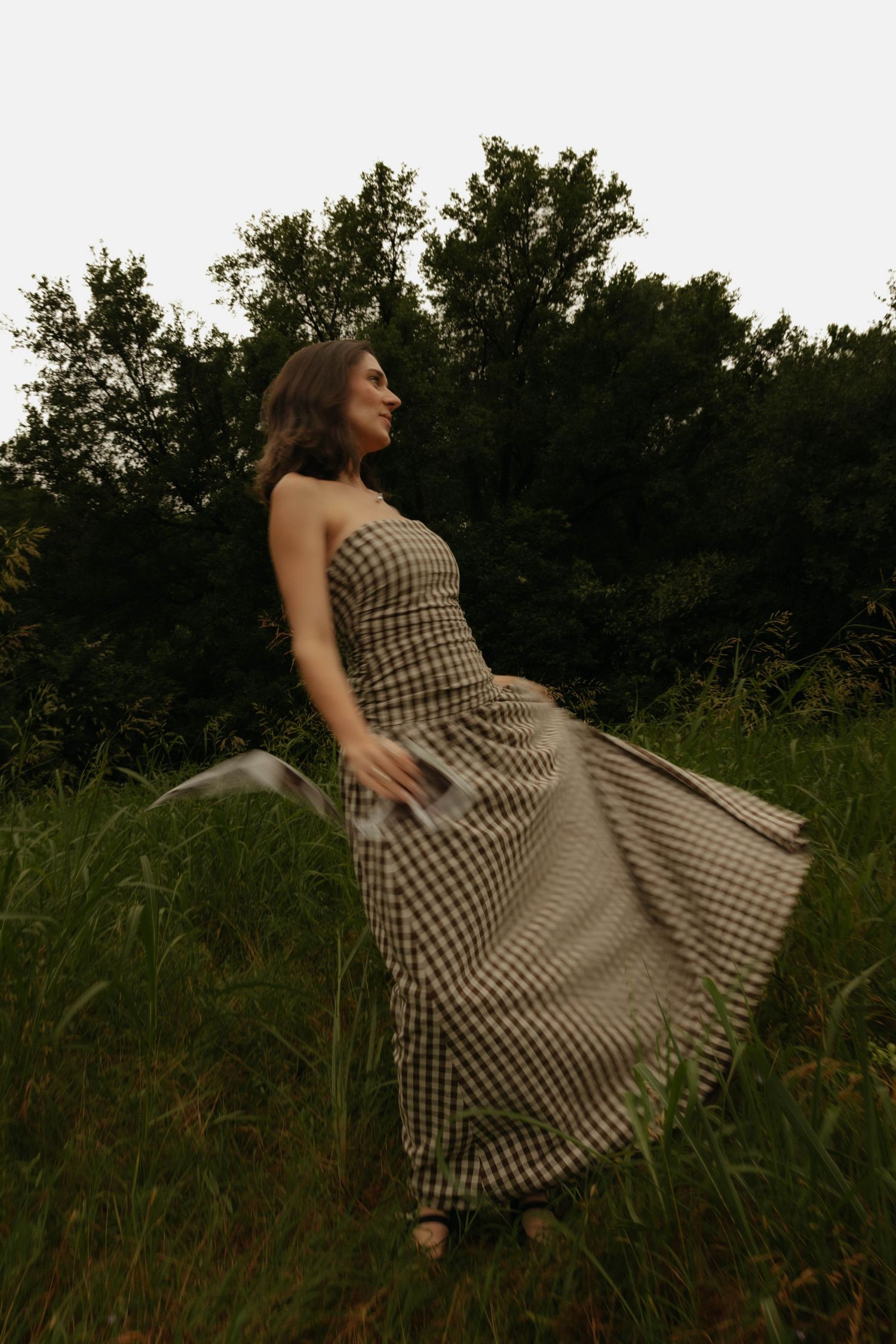 Woman in flowing gingham maxi dress twirling in a green meadow with lush trees in the background.
