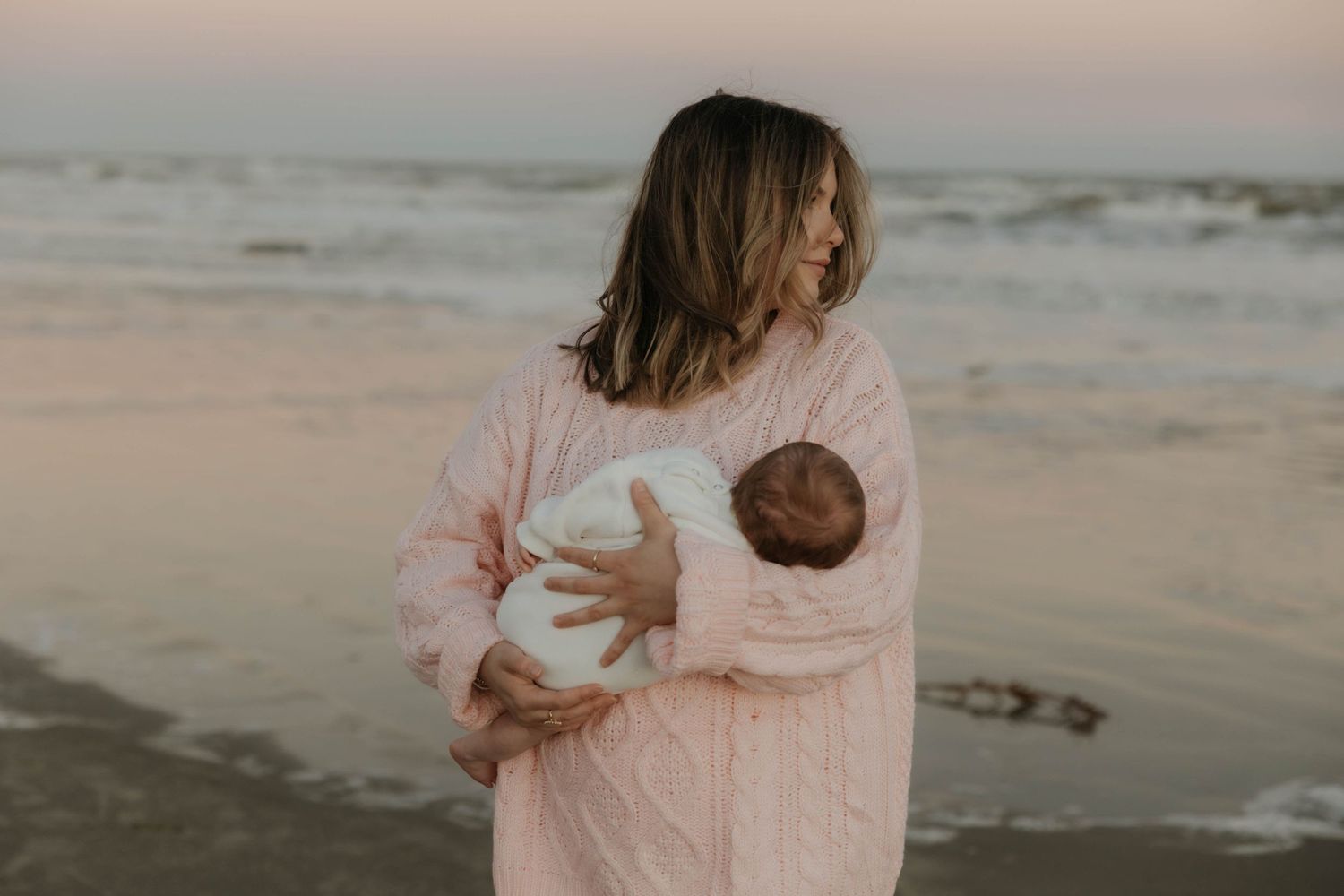 Mother in pink knit sweater cradling newborn baby on a serene beach at sunset.