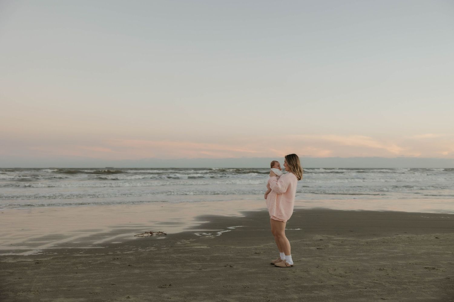 Young mother walking along sandy beach at dusk holding her newborn baby in pink outfit.