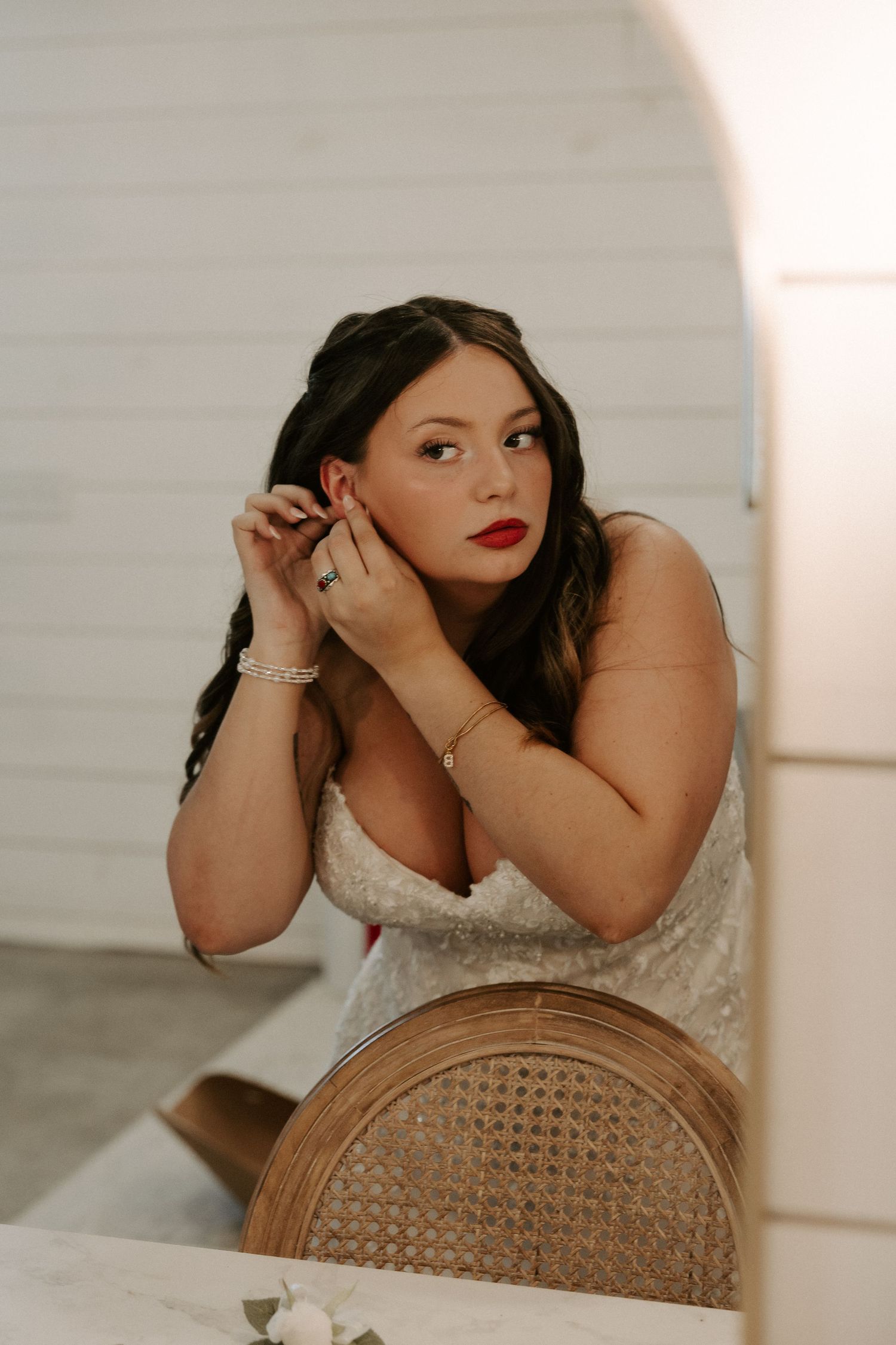 Portrait in white lace dress with red lipstick and dark hair sitting beside a rattan chair in soft lighting.