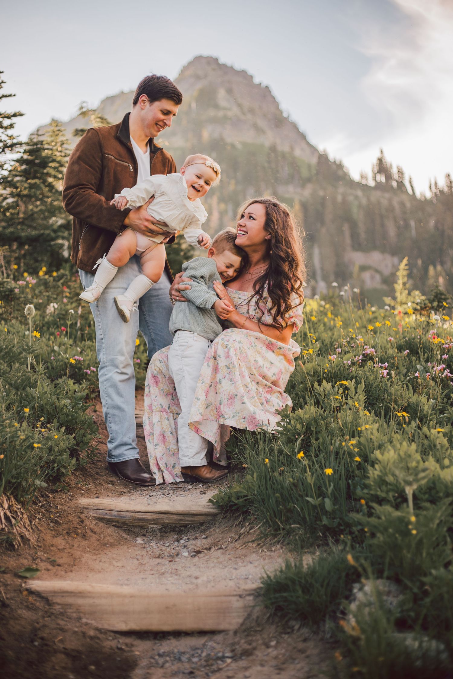 Happy family of four posing on a mountain wildflower trail, parents holding young children with rocky peaks behind.