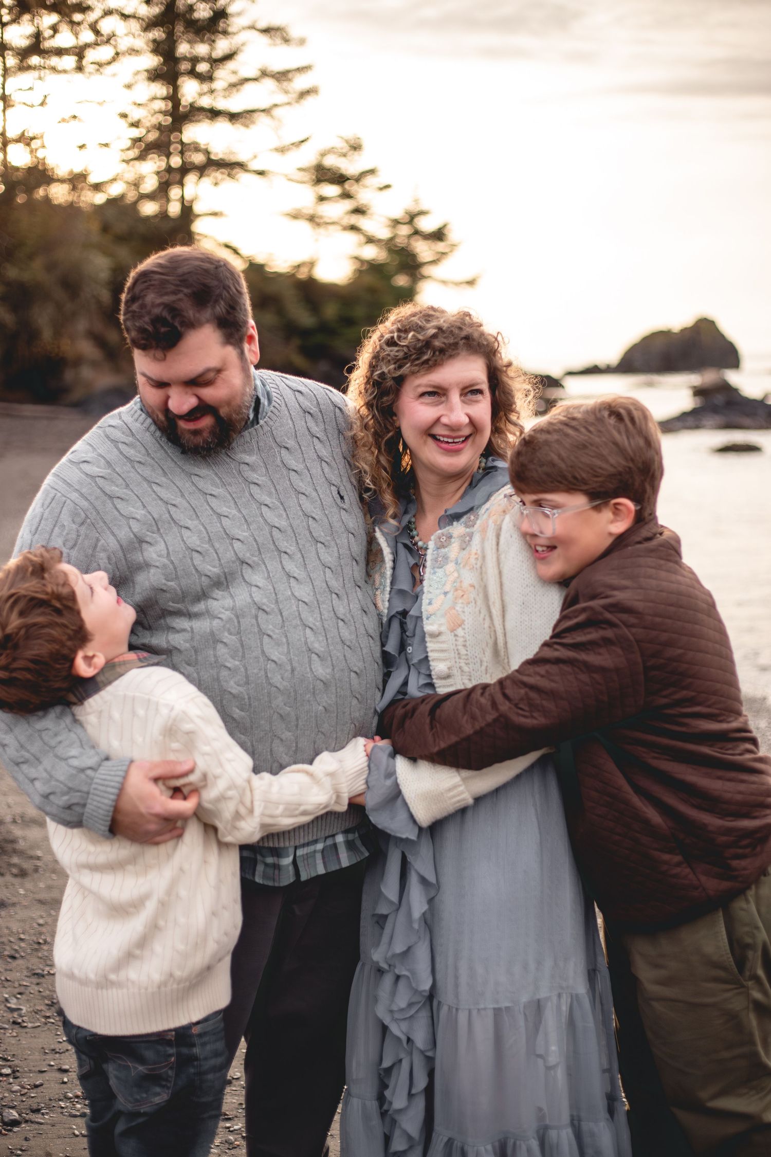 Family of four laughing and embracing on a rocky beach at sunset, with tall trees and ocean rocks in the background.