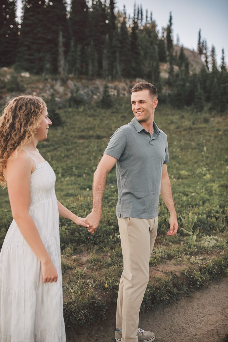 Couple holding hands on a mountain trail, woman in white dress, man in gray polo and khaki pants, with pine trees behind.