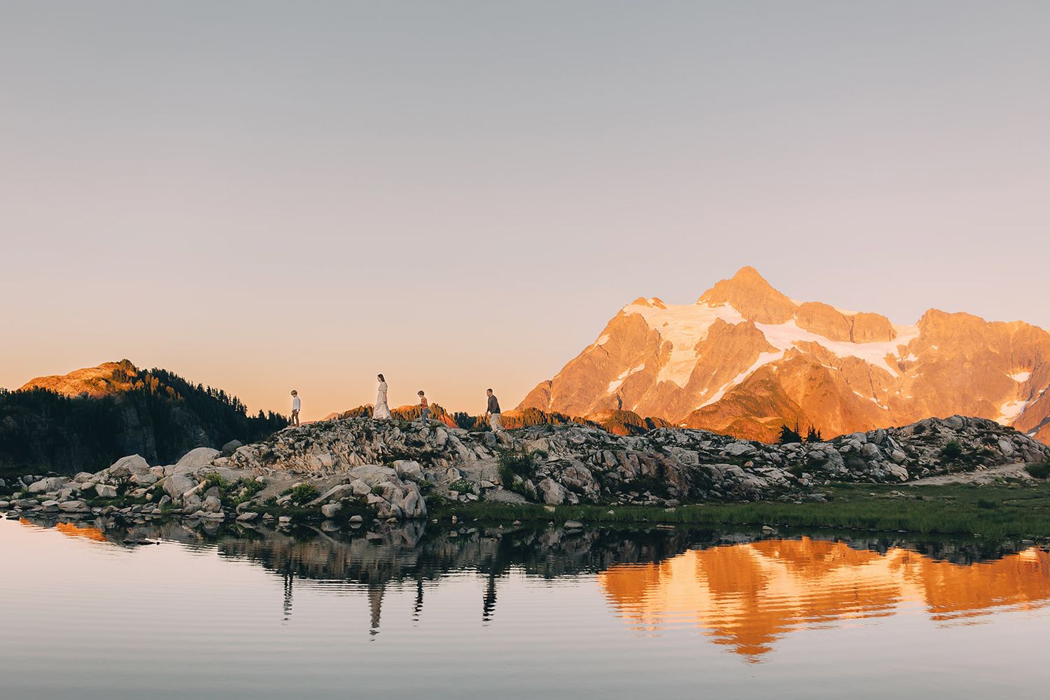 Golden sunset illuminates a snowy mountain peak reflecting in a calm alpine lake with hikers on rocky shoreline.