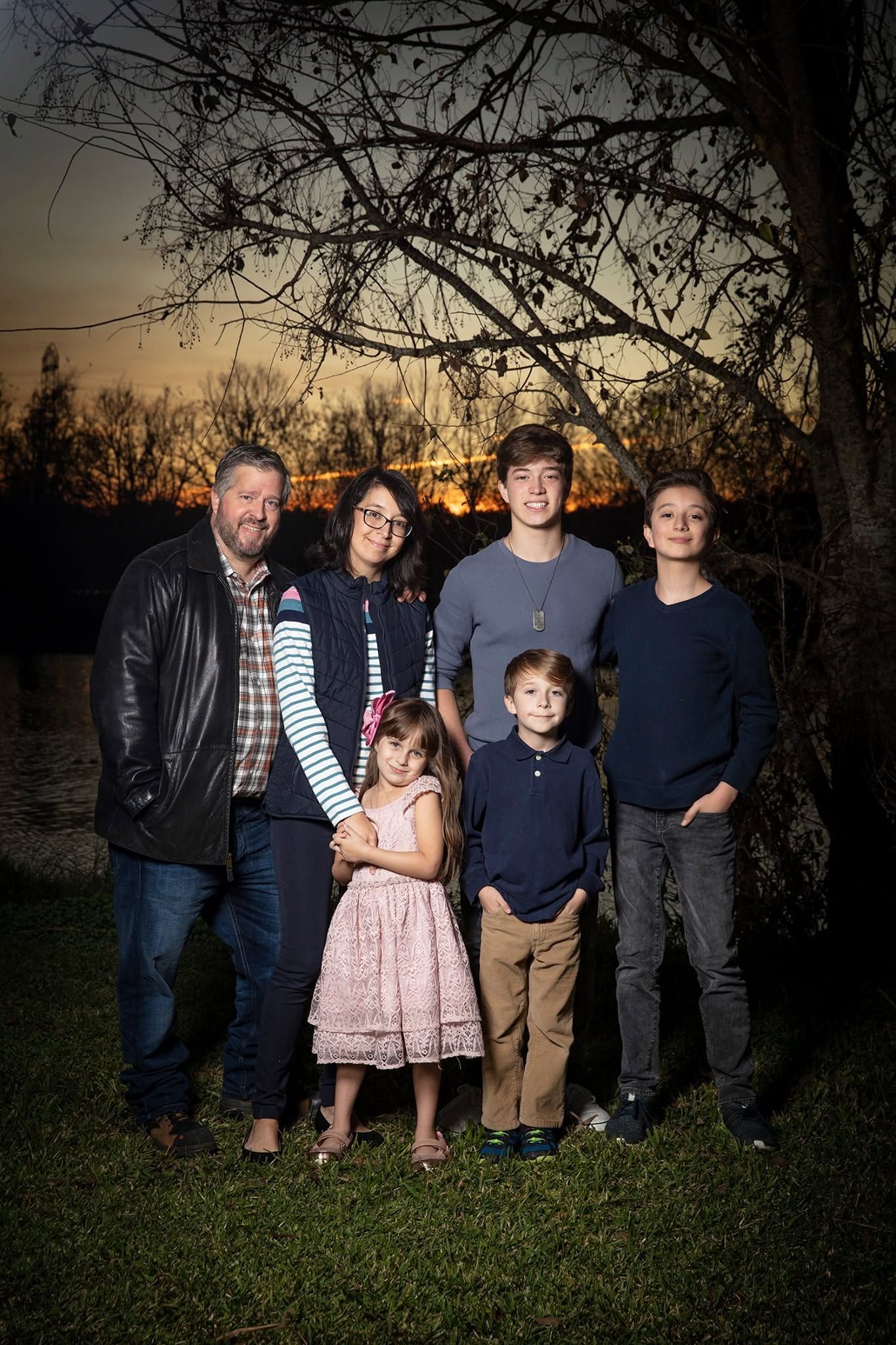 Family of six posing outdoors at sunset near a lake with bare trees in the background.