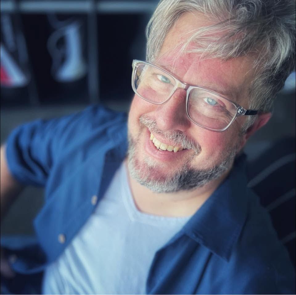 Smiling middle-aged man with gray hair, beard, and glasses wearing a blue shirt, photographed from above indoors.