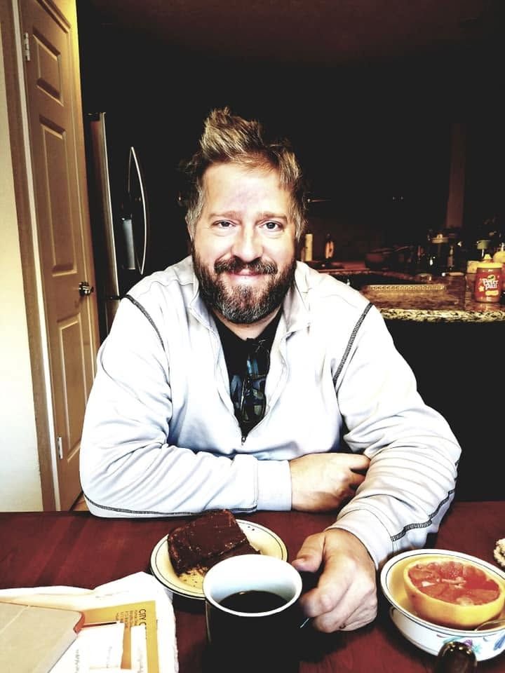 Smiling bearded man sitting at kitchen table with coffee, chocolate cake, and grapefruit breakfast spread.