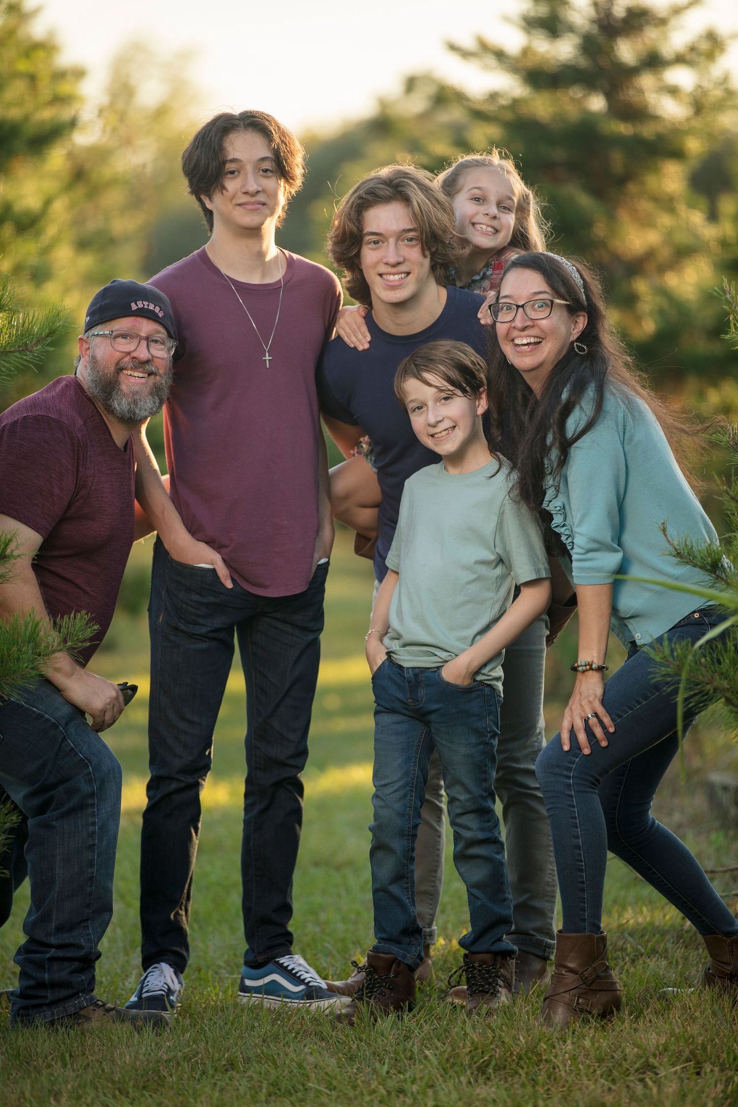 A joyful family of six poses outdoors among pine trees during golden hour, smiling together on green grass.