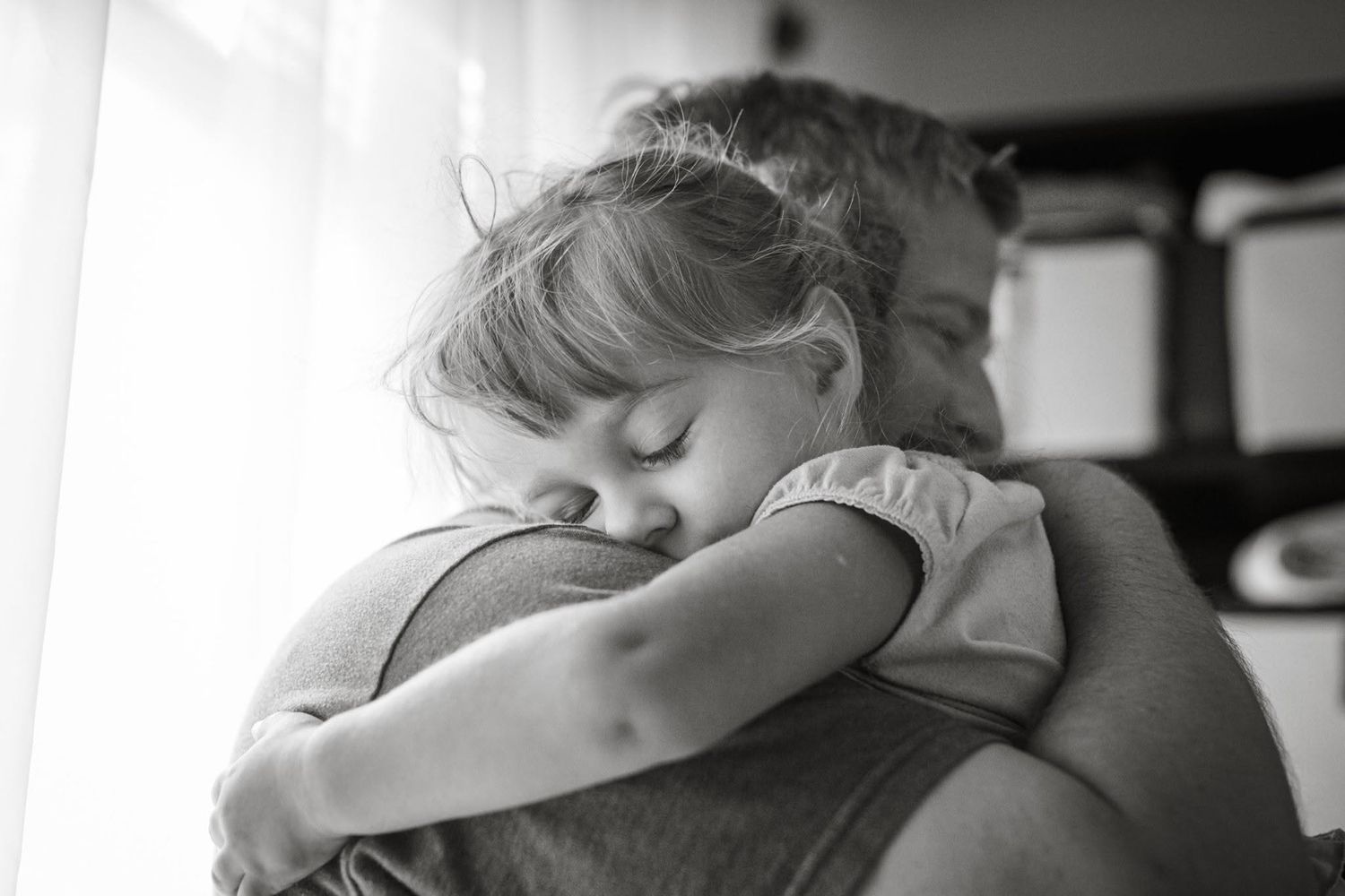 Black and white photo of a young child hugging an adult, eyes closed in a tender embrace near a window.
