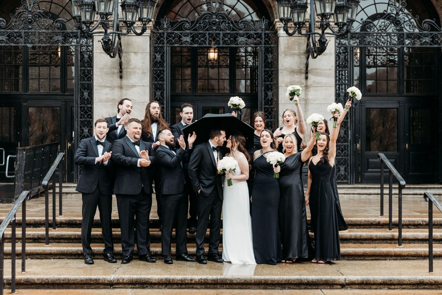 Bride, groom, and bridal party cheering on front steps of Boston Public Library for a group wedding photo on a rainy day in Boston, MA