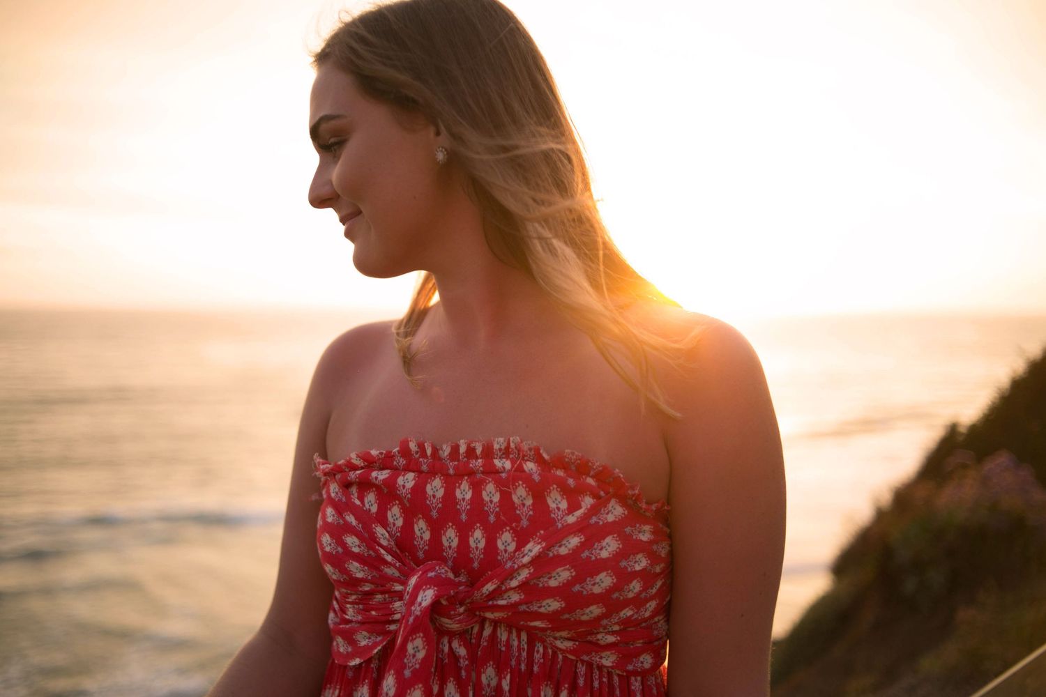Young blonde woman in a red patterned strapless dress gazing at a stunning ocean sunset.