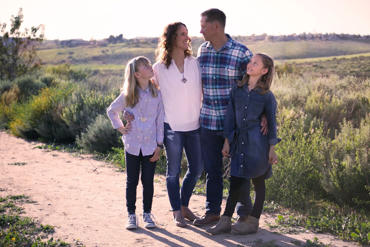 Smiling family of four posing together on a sunny dirt path surrounded by green fields.
