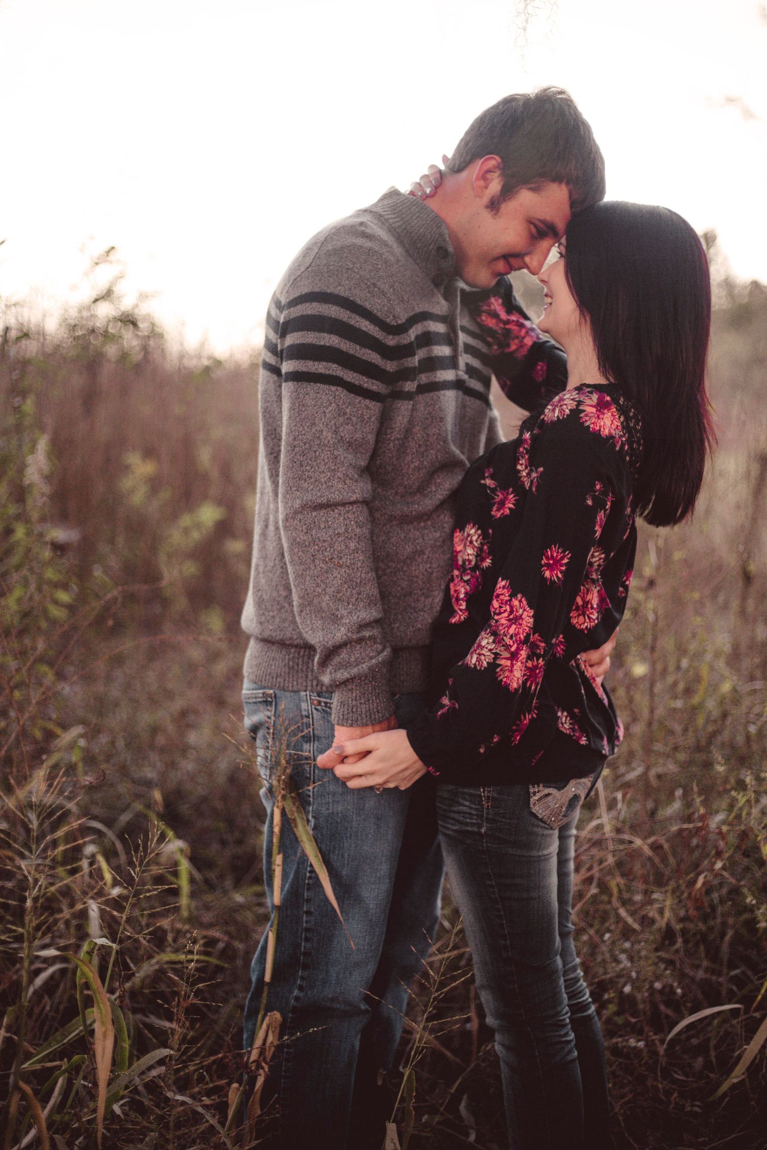 Couple sharing intimate moment in golden field, wearing casual sweaters and jeans at sunset.