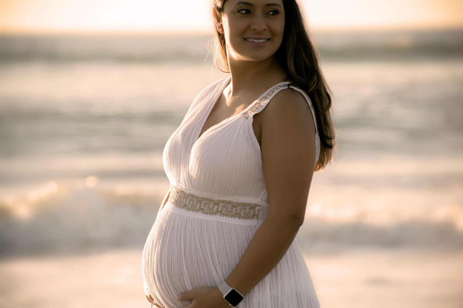 Smiling pregnant woman in white dress holds baby bump on beach at sunset.