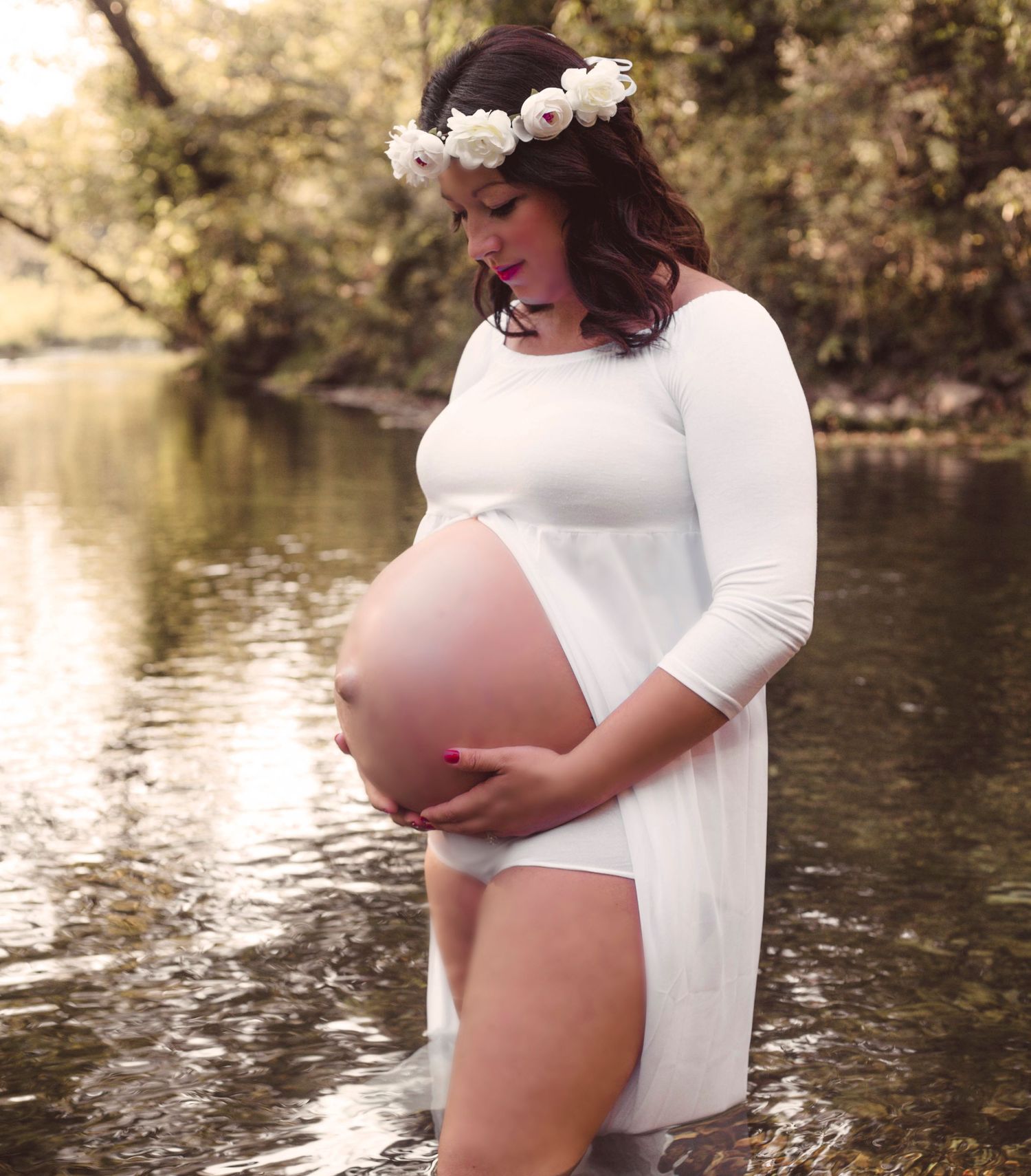 Pregnant woman in white dress and flower crown cradles belly while standing in river.