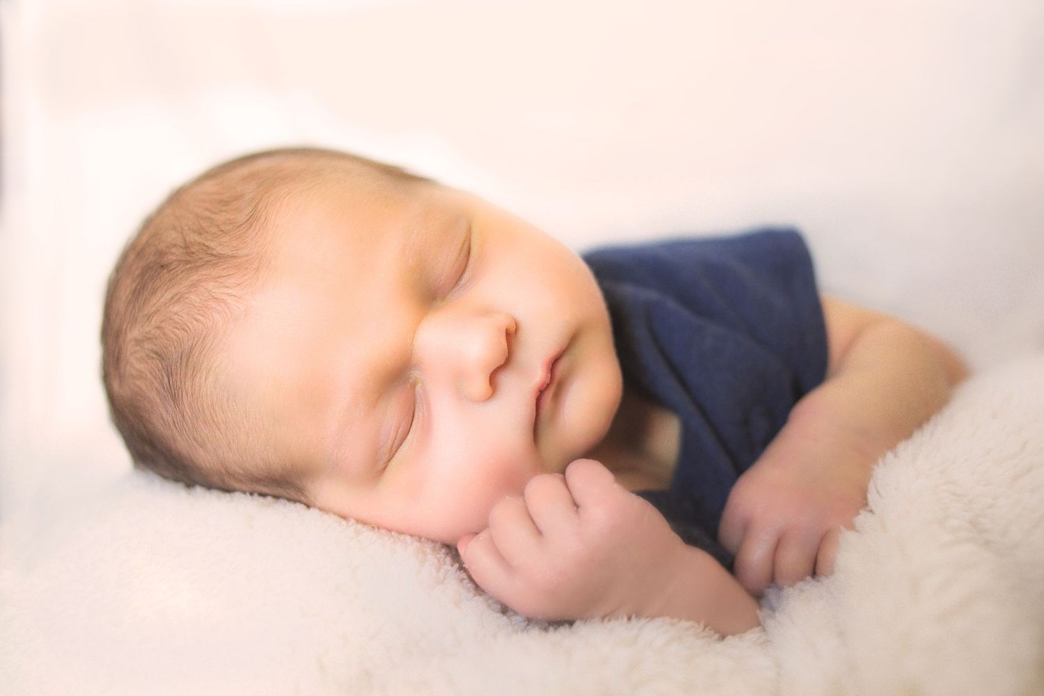 Peaceful newborn baby sleeping on white fluffy blanket wearing a navy blue onesie.