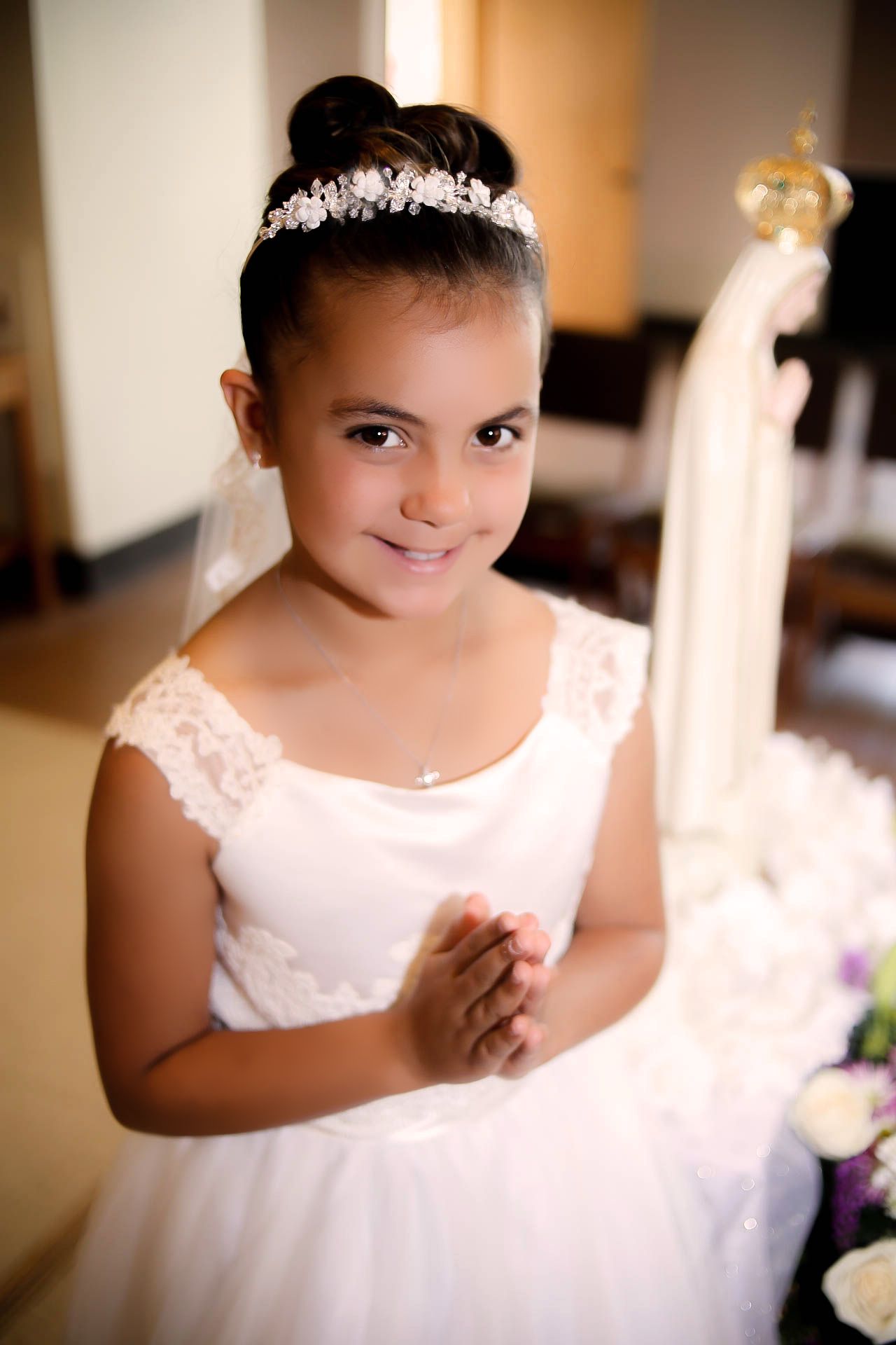 Young girl in white dress and tiara praying at First Communion ceremony beside Virgin Mary statue.