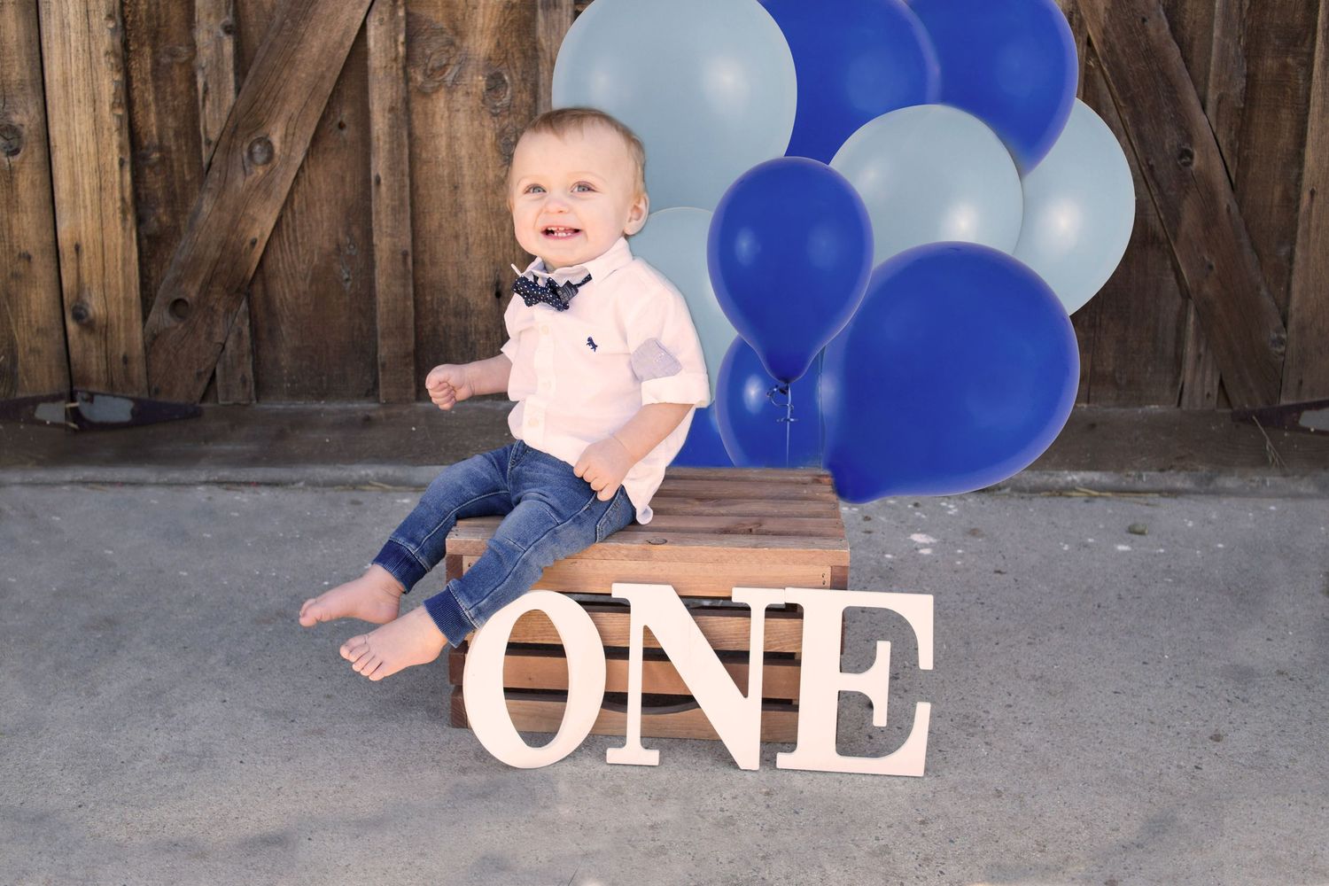 Smiling baby boy celebrating first birthday sitting with ONE sign and blue balloons.
